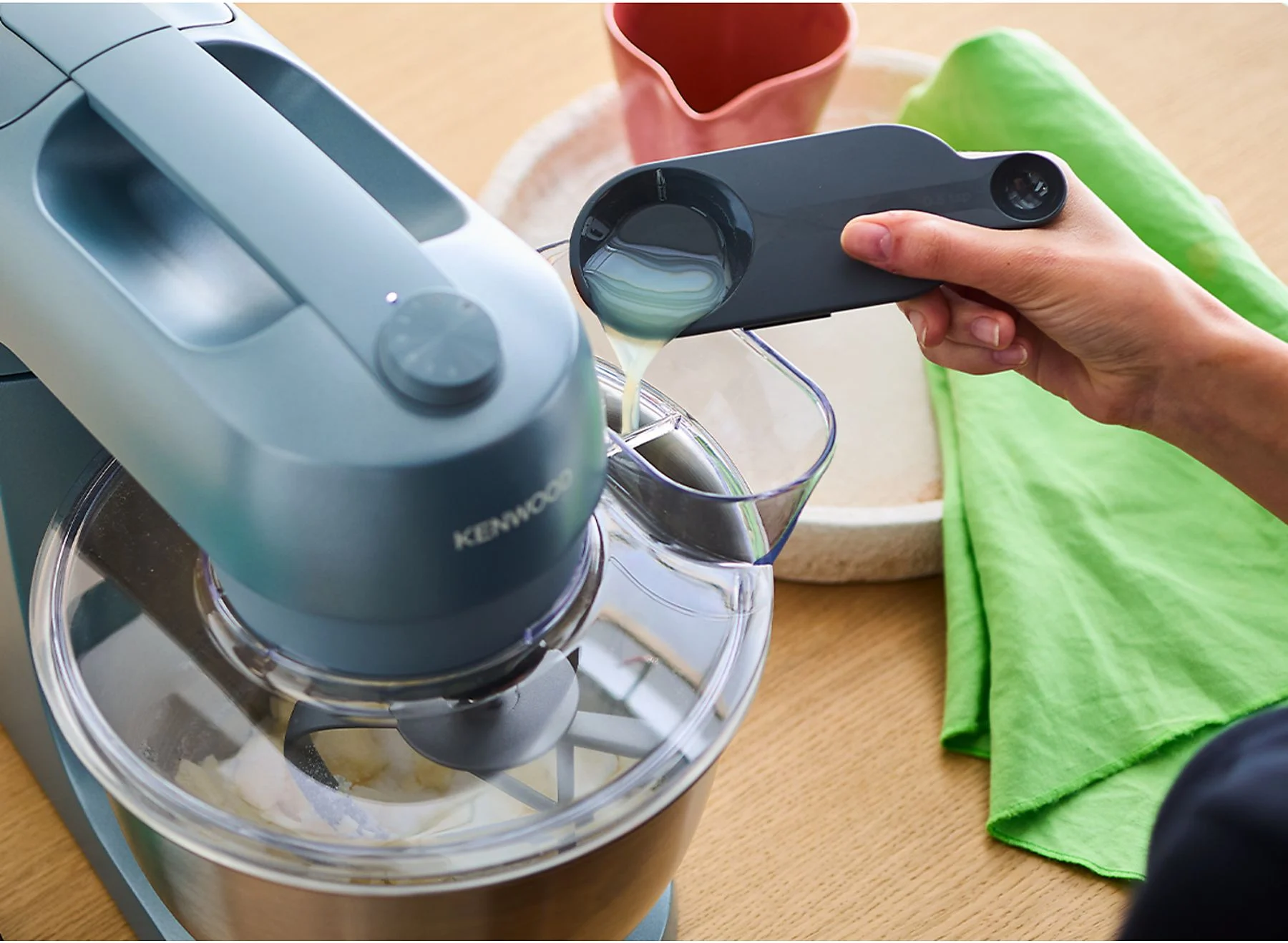 Une femme verse du liquide d'une tasse à mesurer dans un mélangeur. Un bol rose et un chiffon vert sont visibles.