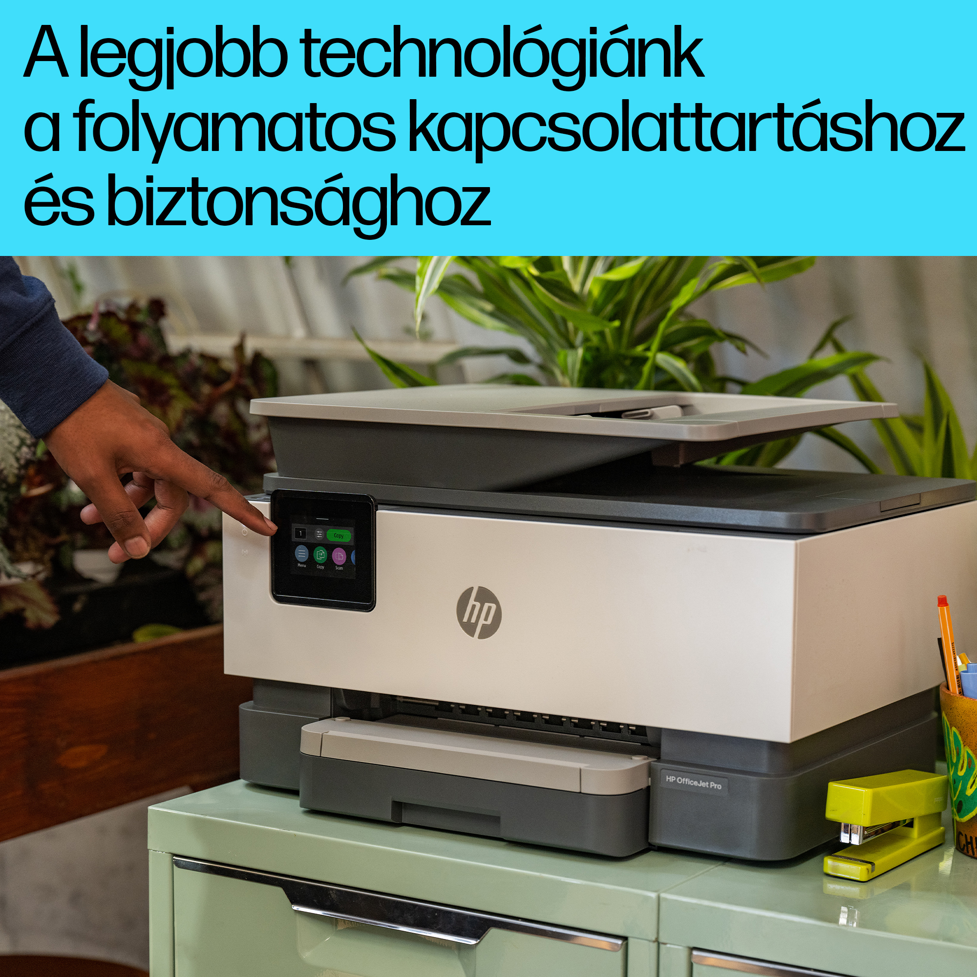 Egy személy megérinti a nyomtató képernyőjét. A nyomtató egy zöld szekrényen van. A szöveg a kép tetején található.