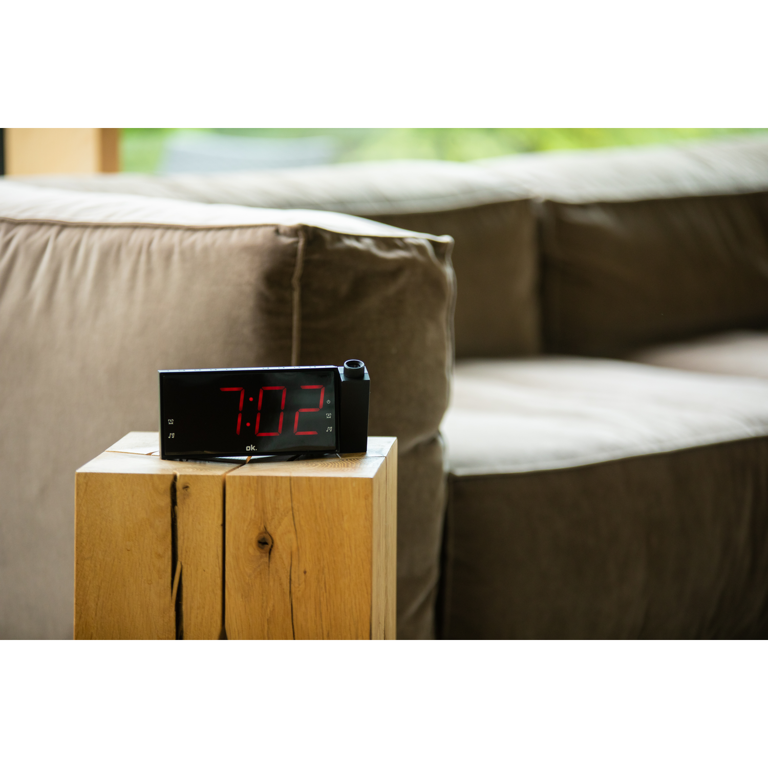 Black digital clock with red digits, on a wooden block. Sofa in the background.