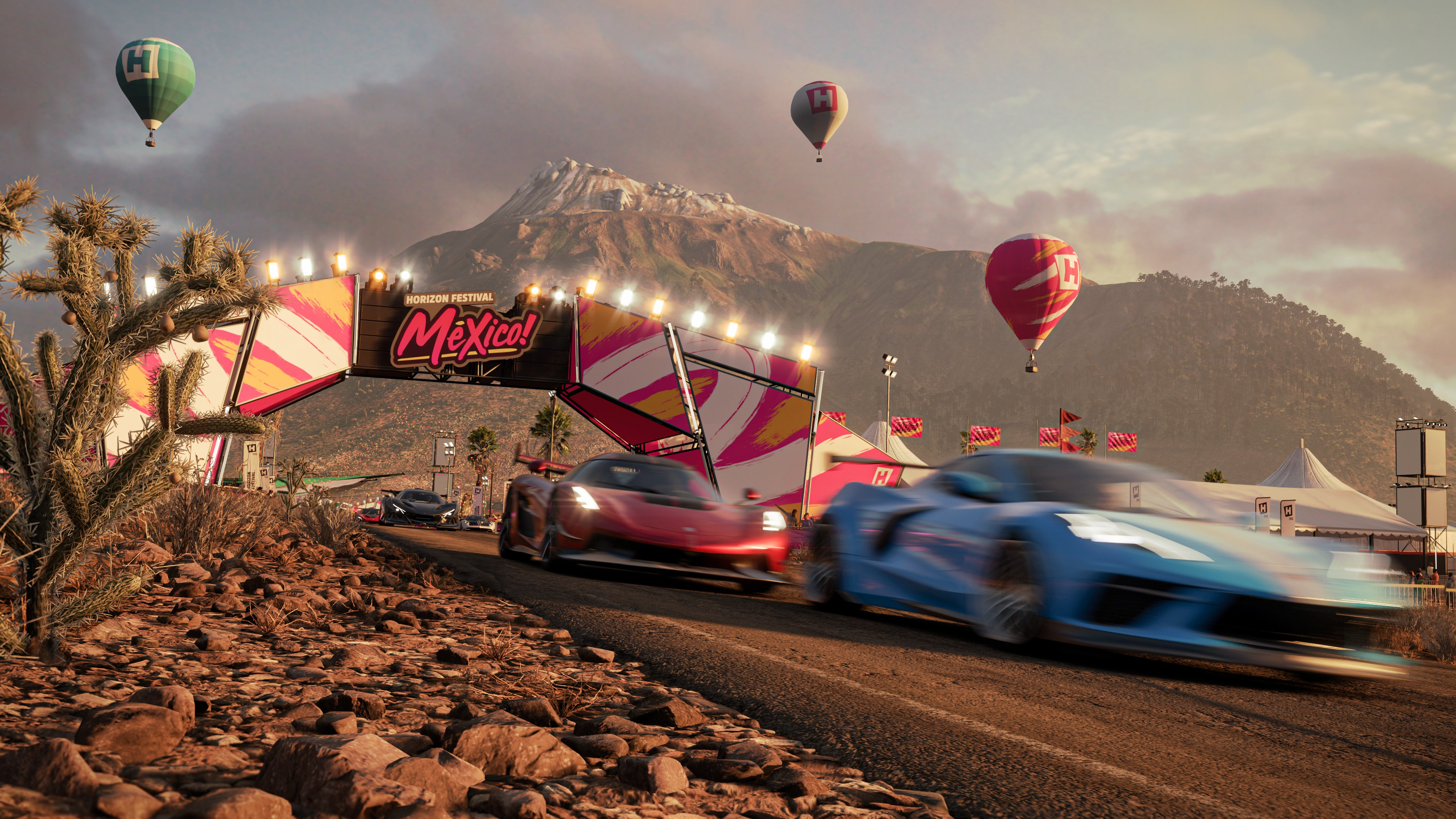 Rennszene: Autos rasen unter einem Festivalbogen mit Heißluftballons am Himmel und einem Berg im Hintergrund.