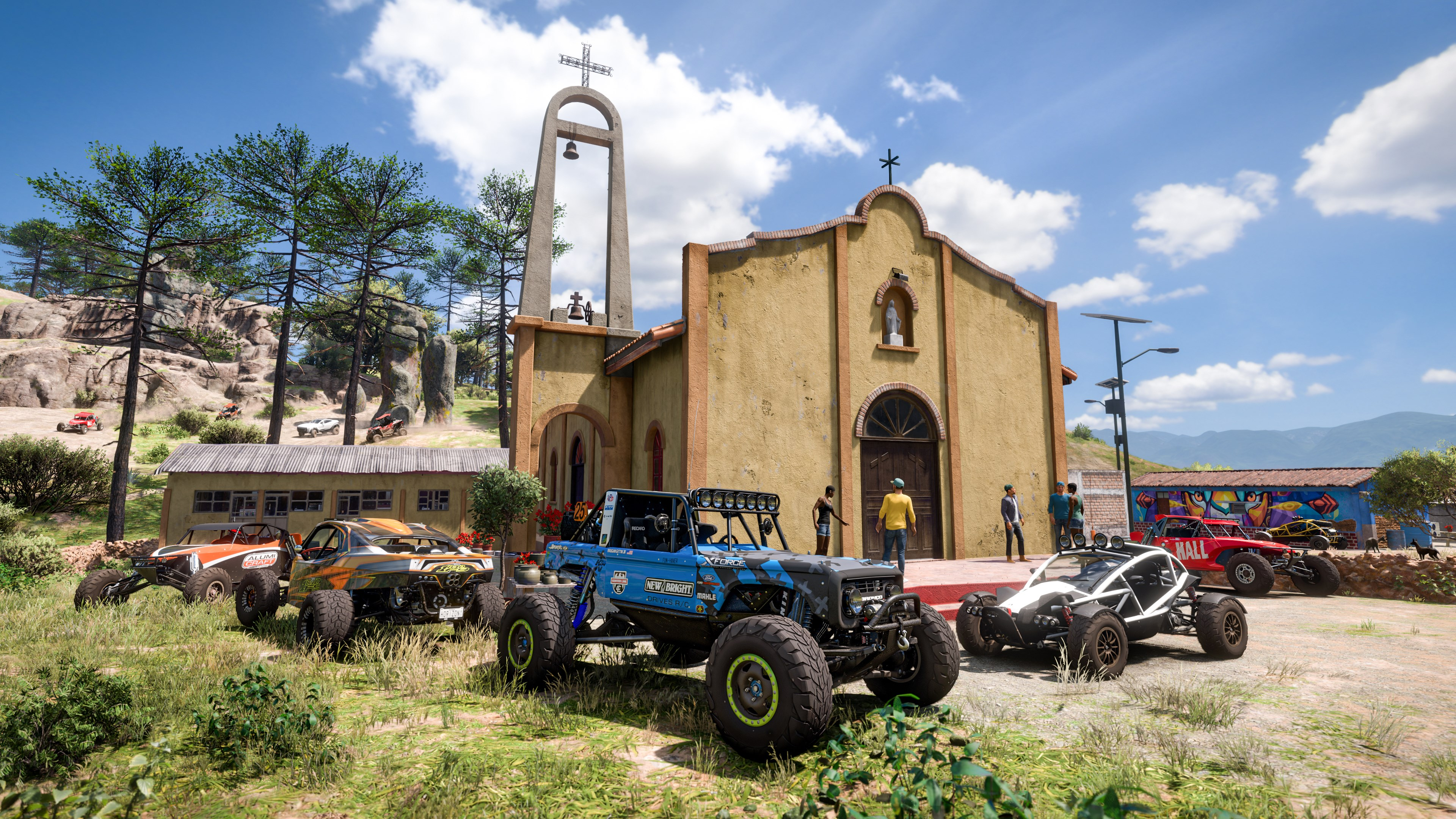 Geländewagen parken vor einer Kirche. Menschen stehen in der Nähe. Bäume und ein Berg sind im Hintergrund unter blauem Himmel.