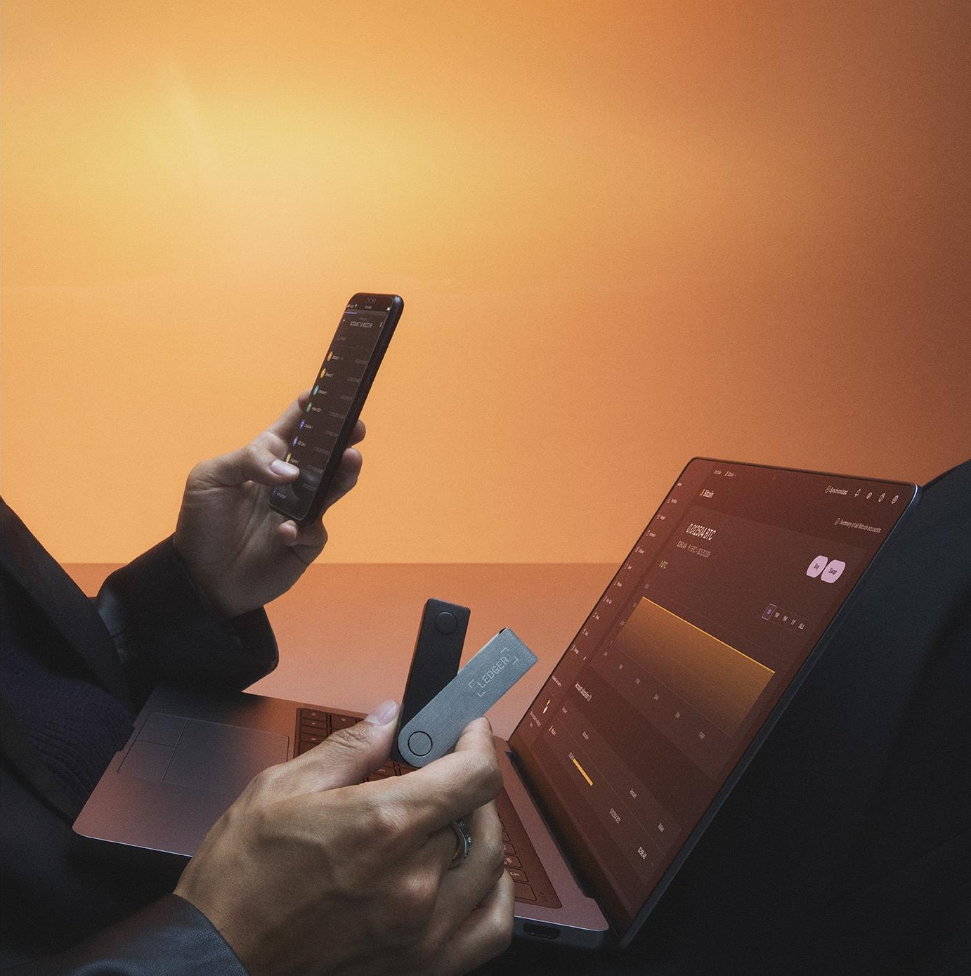 Una persona tiene in mano un telefono e due portafogli hardware crittografici vicino a un laptop che mostra un grafico bitcoin, su sfondo arancione.