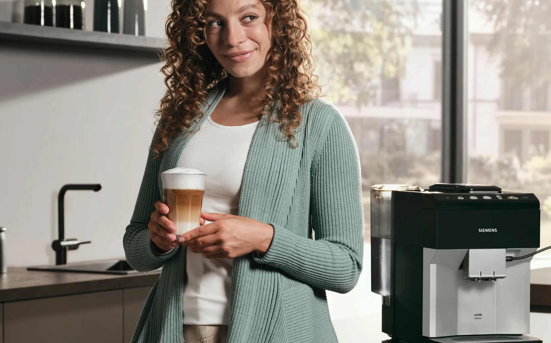 Kobieta trzymająca latte, stojąca obok ekspresu do kawy. Tło to kuchnia.