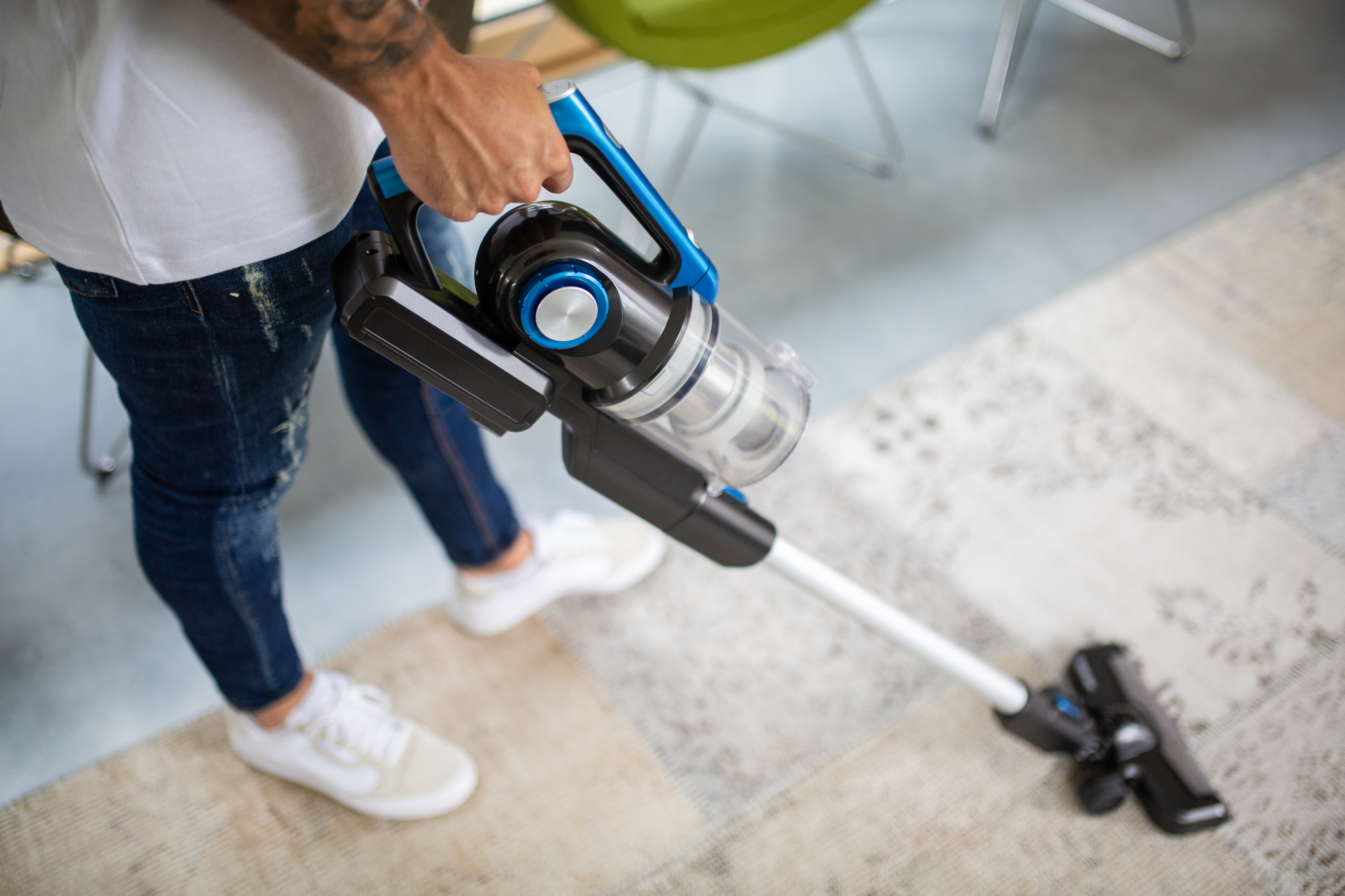Mężczyzna trzyma bezprzewodowy odkurzacz, odkurzając dywan. Odkurzacz jest czarny i niebieski.
