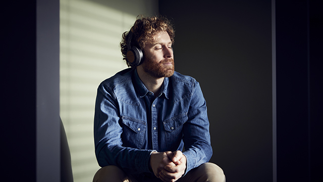 Mężczyzna ze słuchawkami, zamknięte oczy. Siedzi w świetle słonecznym. Ciemne tło.