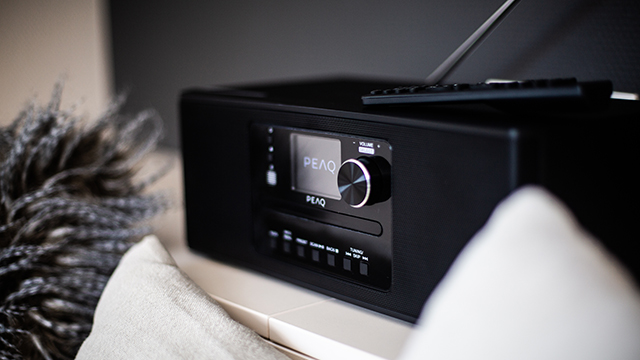 Czarny radioodtwarzacz PEAQ z pilotem i białą poduszką w tle.