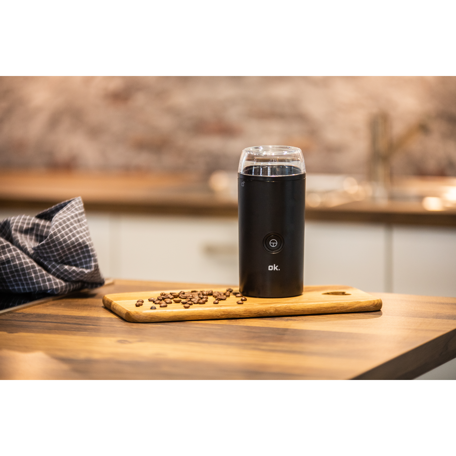 A black coffee grinder with coffee beans on a wooden cutting board. The background is a kitchen counter.