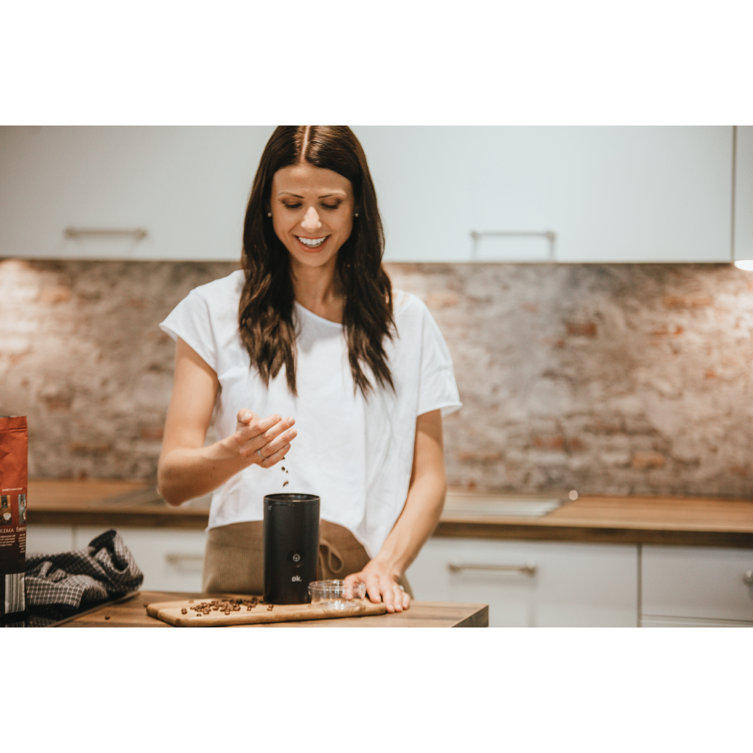 A woman smiles, holding coffee beans and pouring them into a black grinder on a wooden board.