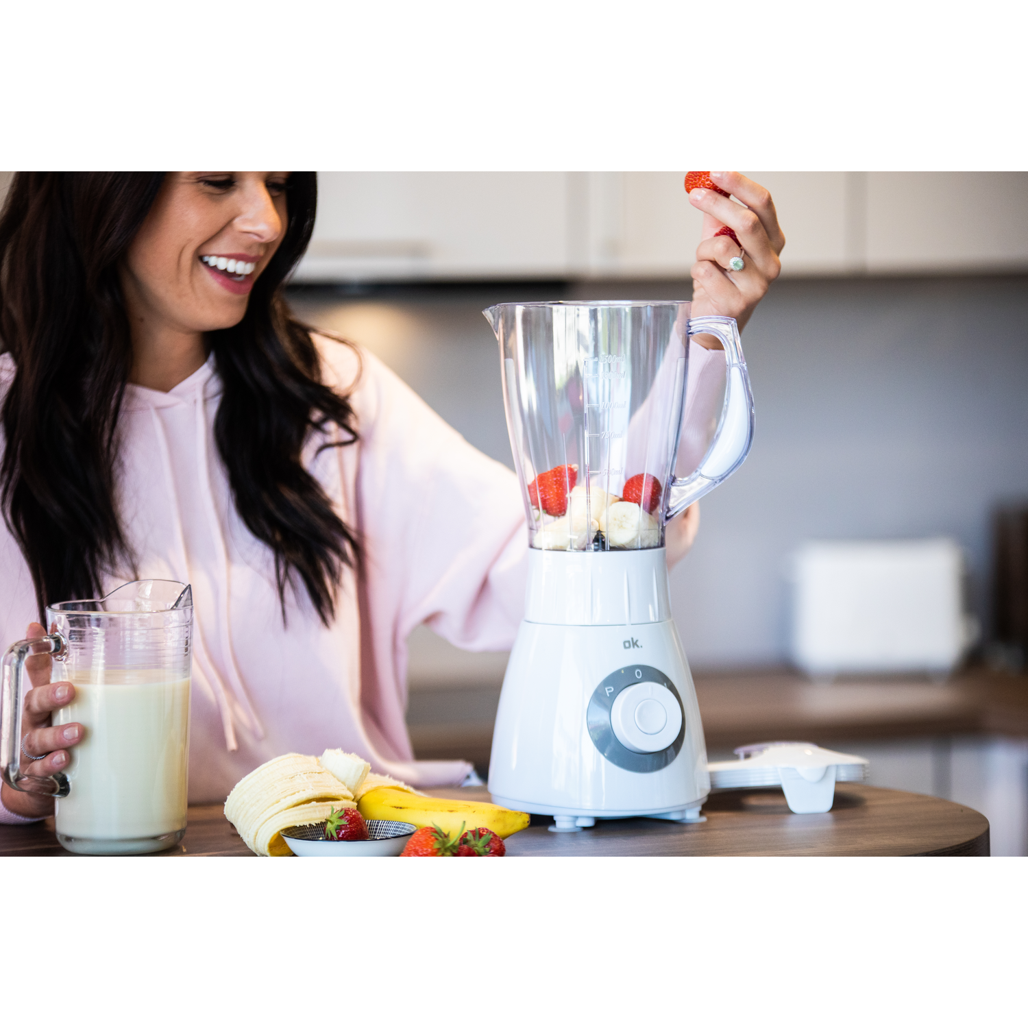 Vrouw lachend, voegt aardbei toe aan blender. Blender met fruit erin. Melkkan en bananenschijfjes in de buurt.