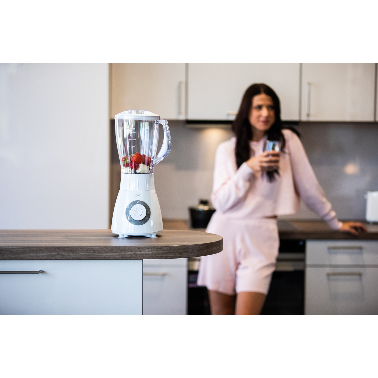 Een vrouw staat bij een blender op een aanrecht met fruit en een keukenachtergrond.