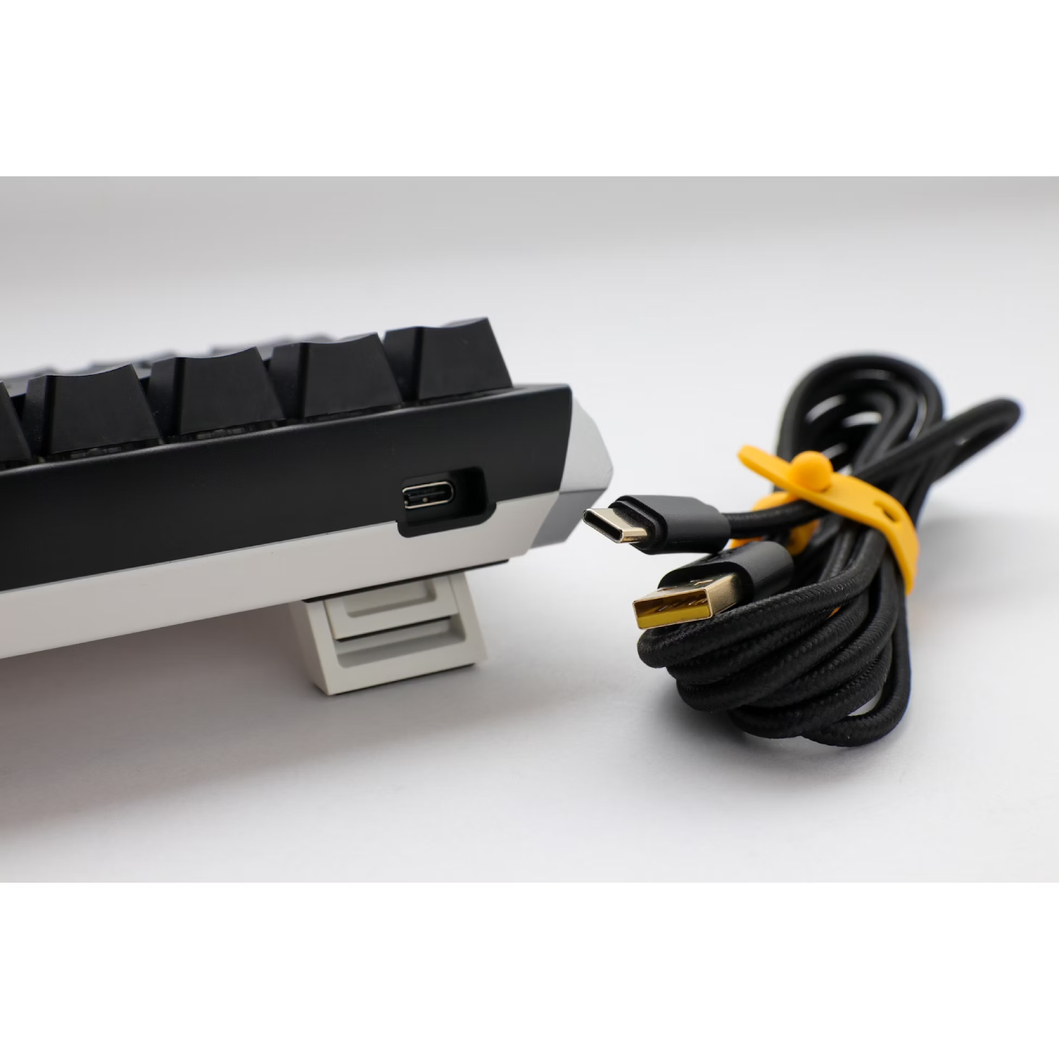 Un teclado negro con cable USB-C y brida amarilla, todo sobre fondo blanco.