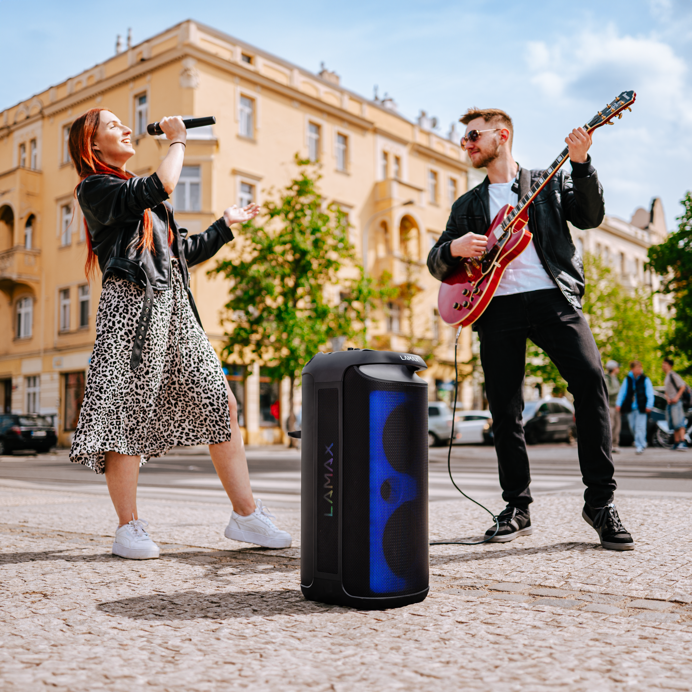 Egy nő és egy férfi énekelnek és gitároznak egy városi téren egy hordozható hangszóró mellett.