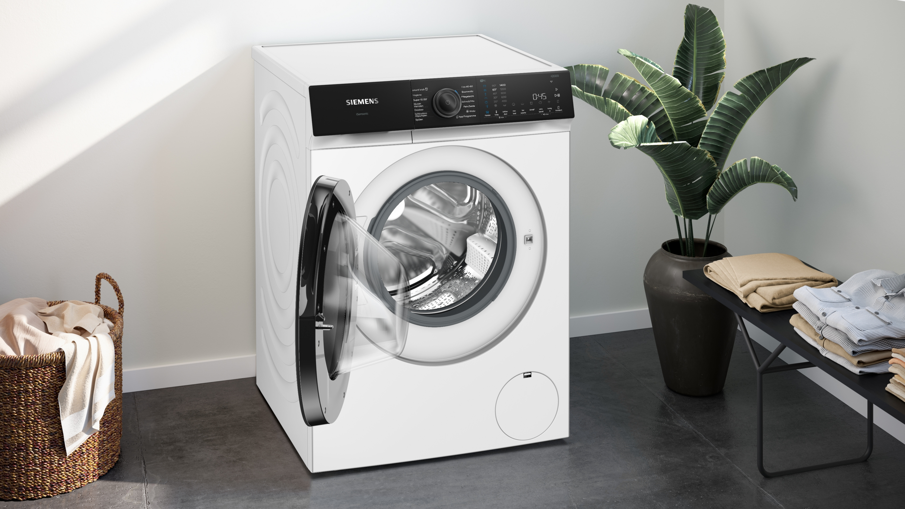 Eine weiße Waschmaschine mit offener Tür, neben einer Pflanze und einem Korb.
