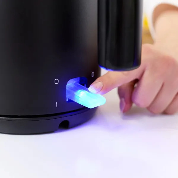 Palec osoby naciska podświetlany na niebiesko przełącznik na czarnym czajniku. Czajnik stoi na białej powierzchni.