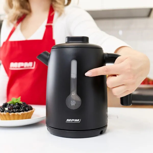 Osoba wskazuje na czajnik elektryczny w kolorze czarnym. W pobliżu czajnika talerz z małym deserem.