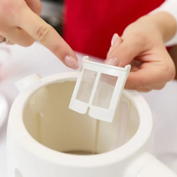 Dłoń trzyma biały filtr do herbaty nad białym czajnikiem, wskazując.