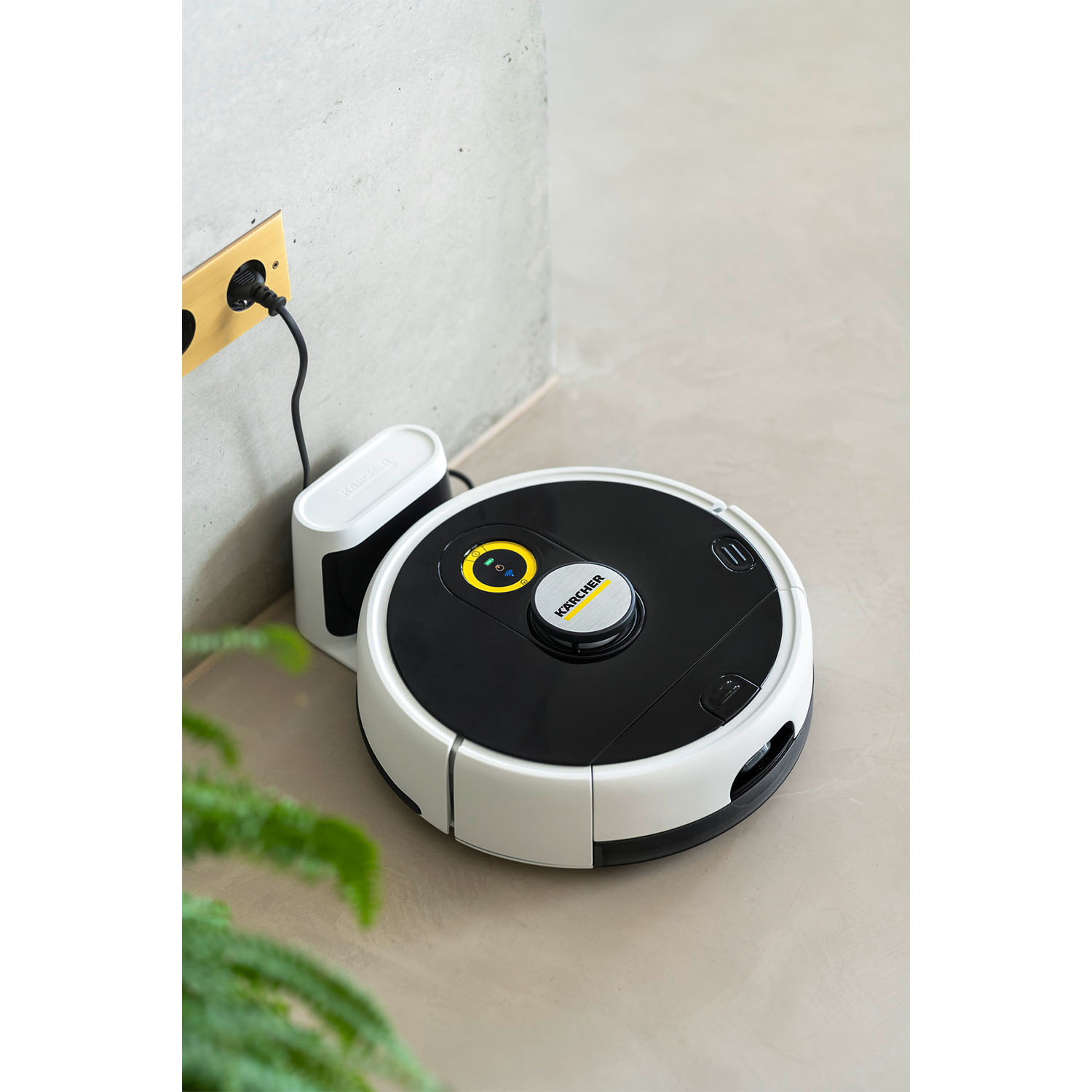 Aspirapolvere robot sul pavimento vicino a una stazione di ricarica e una pianta.