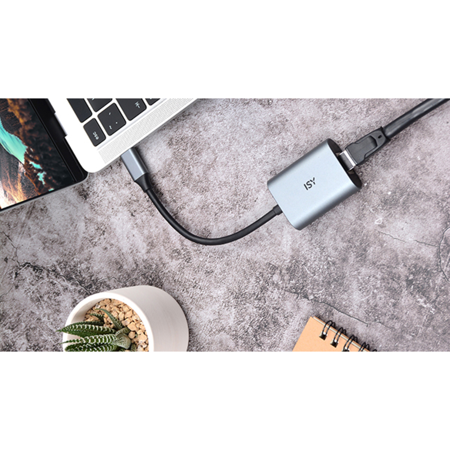 Ordinateur portable connecté à un adaptateur gris via un câble noir. Une petite plante et un carnet sont présents.