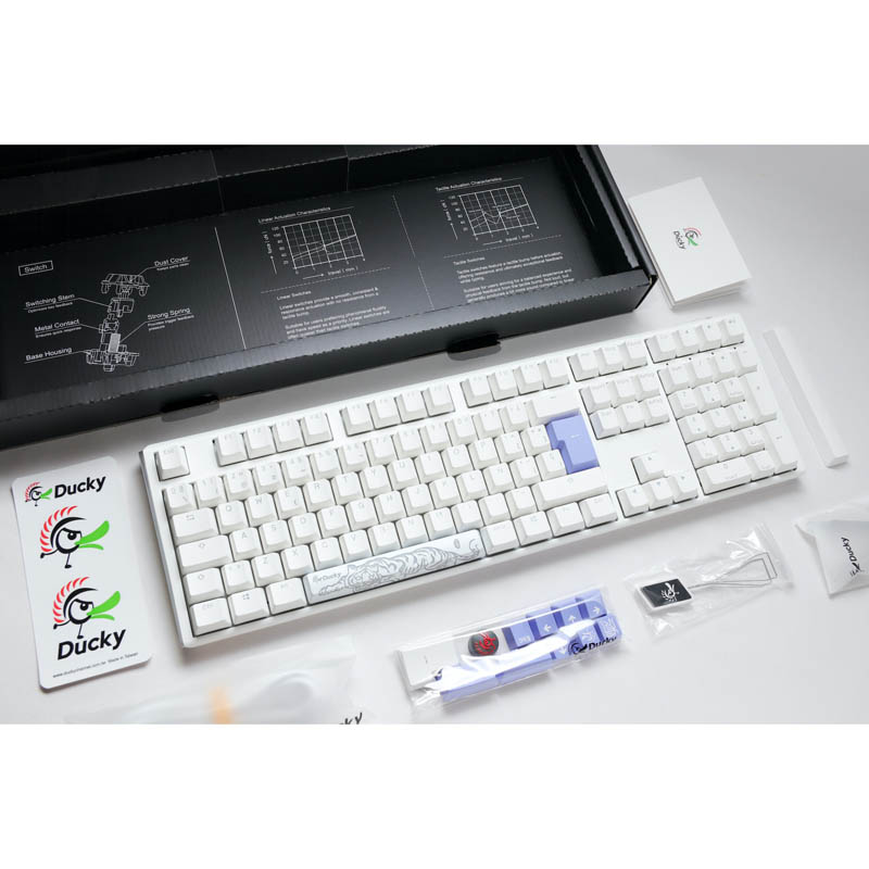 Un teclado mecánico blanco con teclas azules, accesorios y una caja negra. Vista superior.