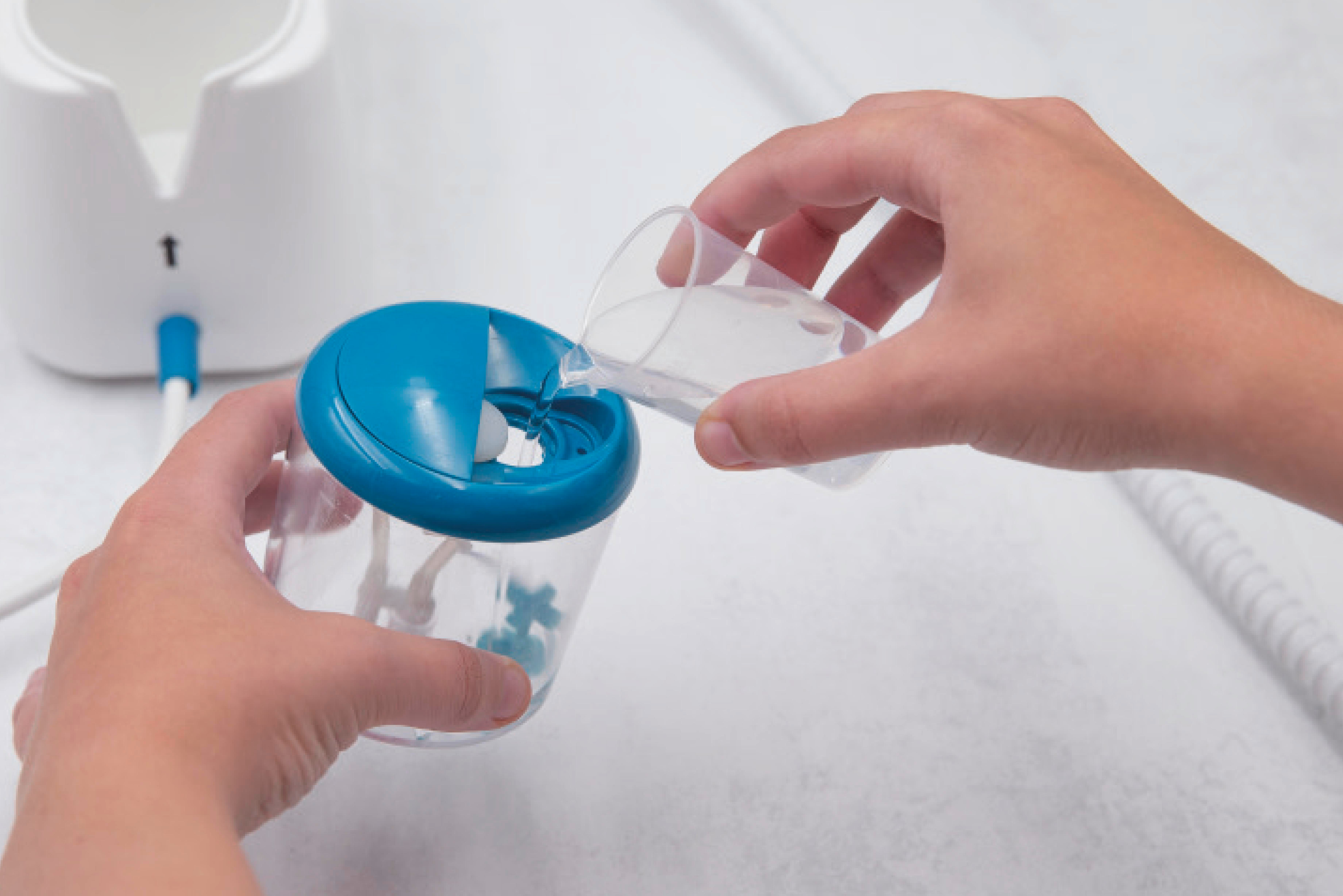Hände gießen Wasser in einen blauen Behälter. Links ist ein weißes Gerät mit langem weißem Kabel.