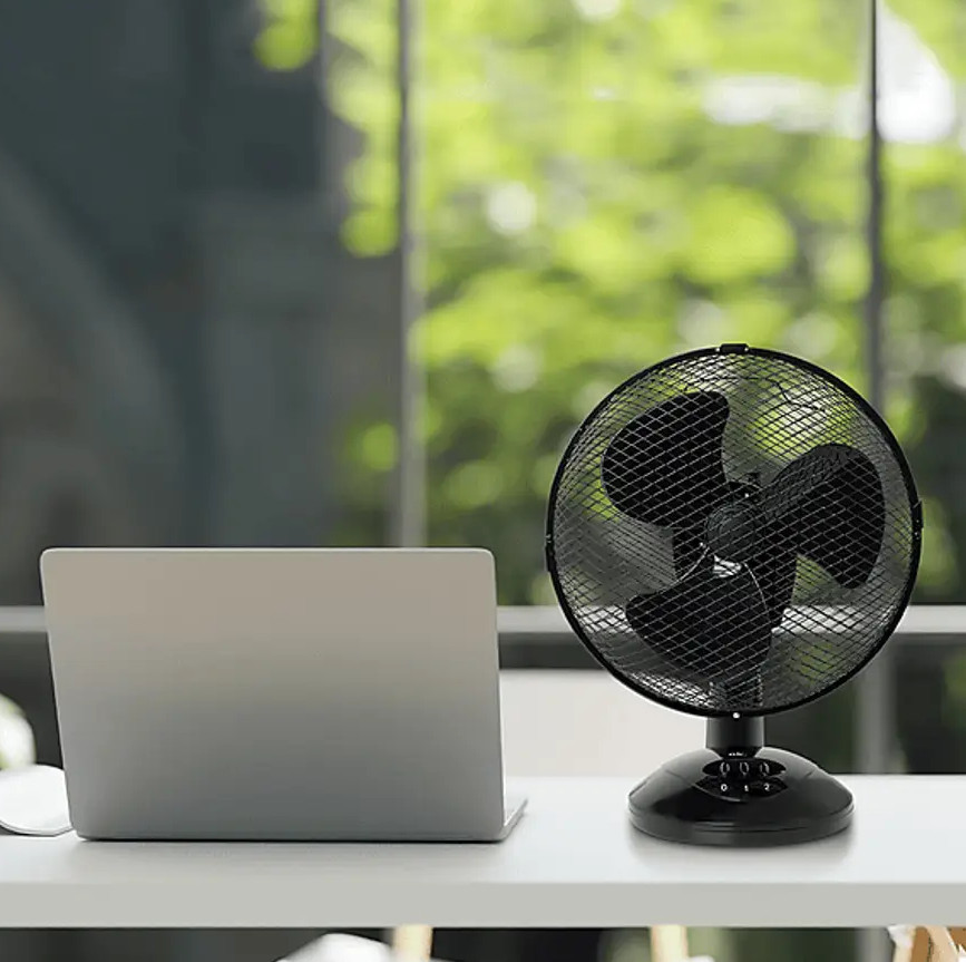 Czarny wentylator biurkowy obok laptopa na białym biurku. Zielone liście widoczne przez okno w tle.