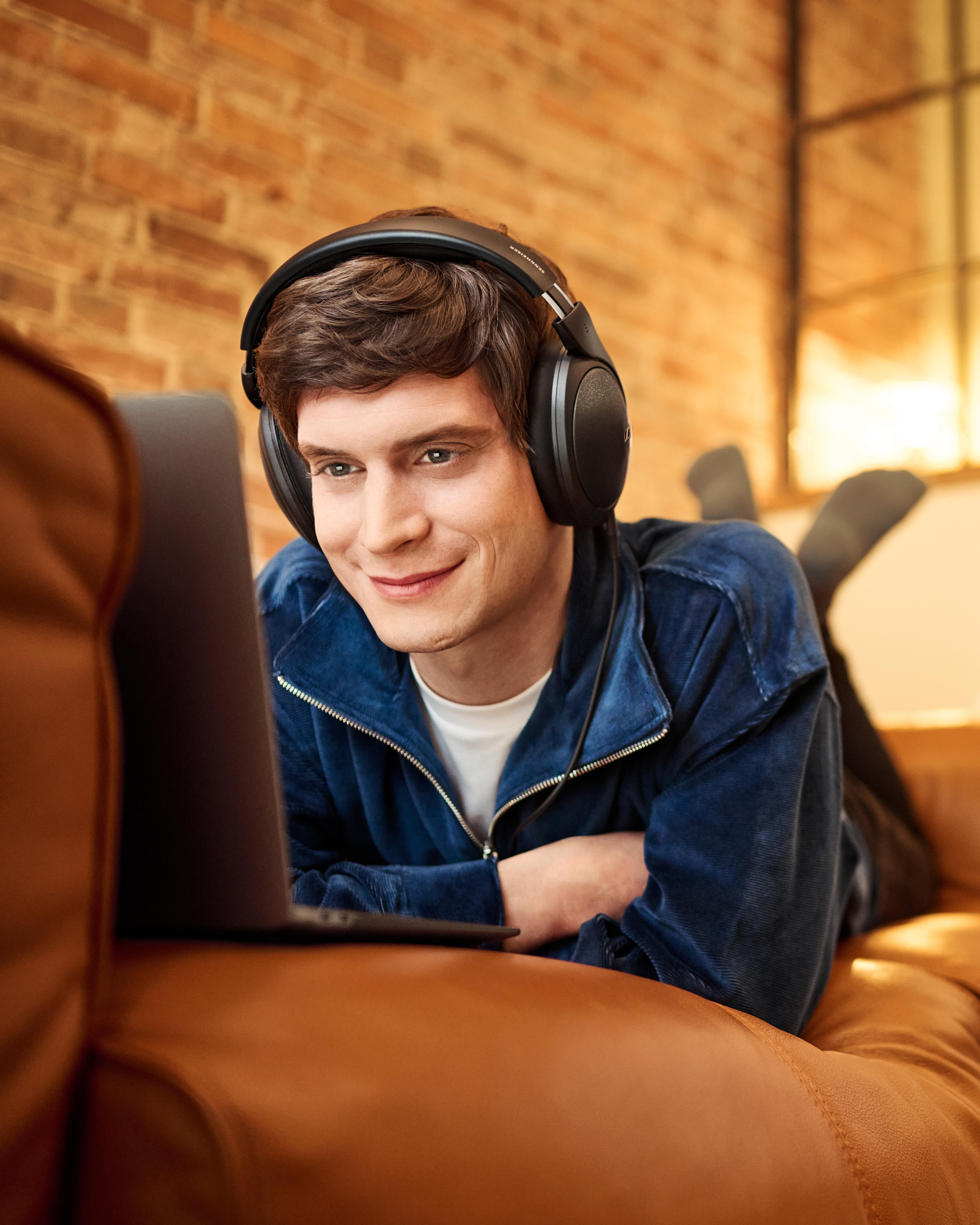Mann liegt auf Couch, trägt Kopfhörer, schaut auf Laptop.
