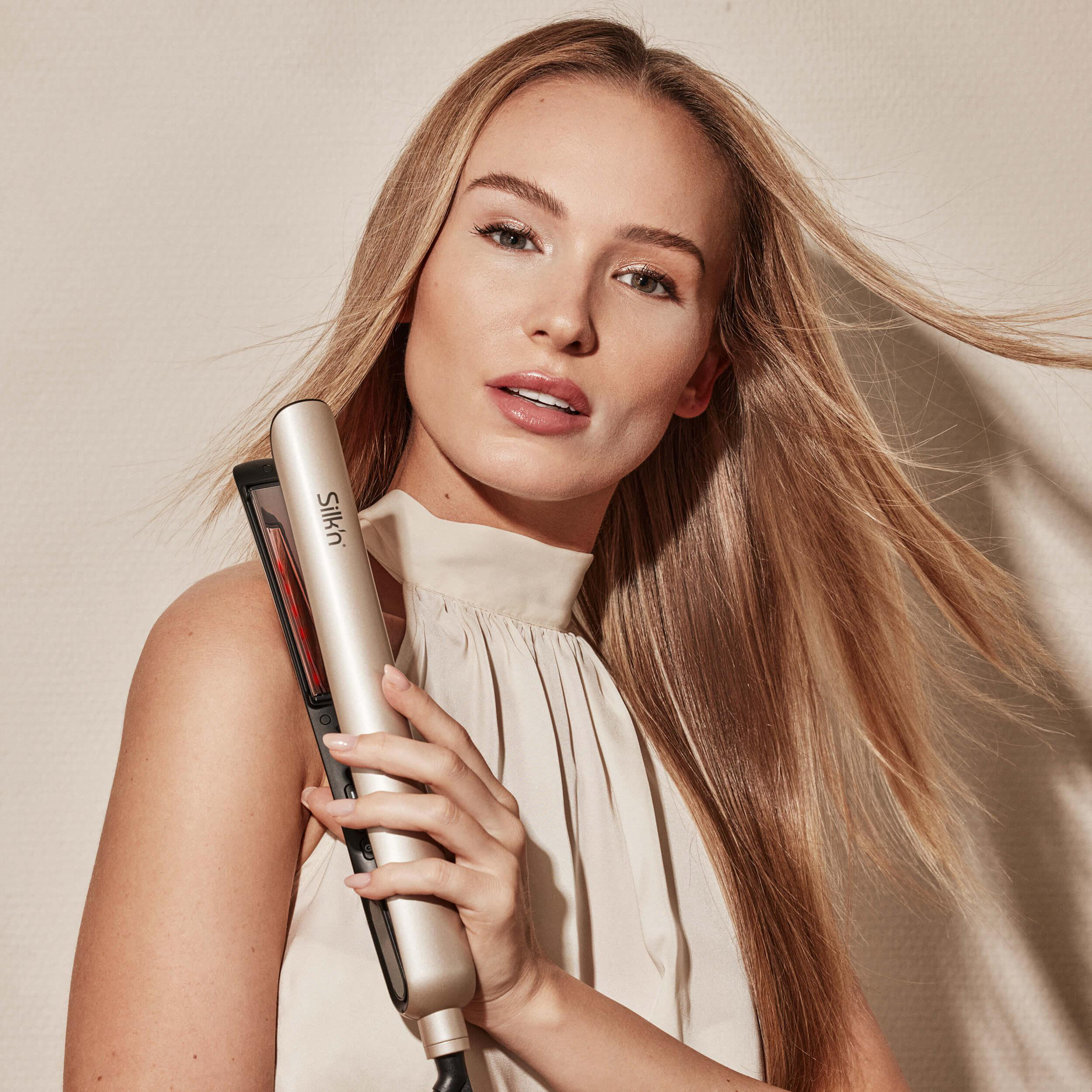 Eine Frau hält einen Haarglätter. Sie hat lange blonde Haare und blickt in die Kamera.