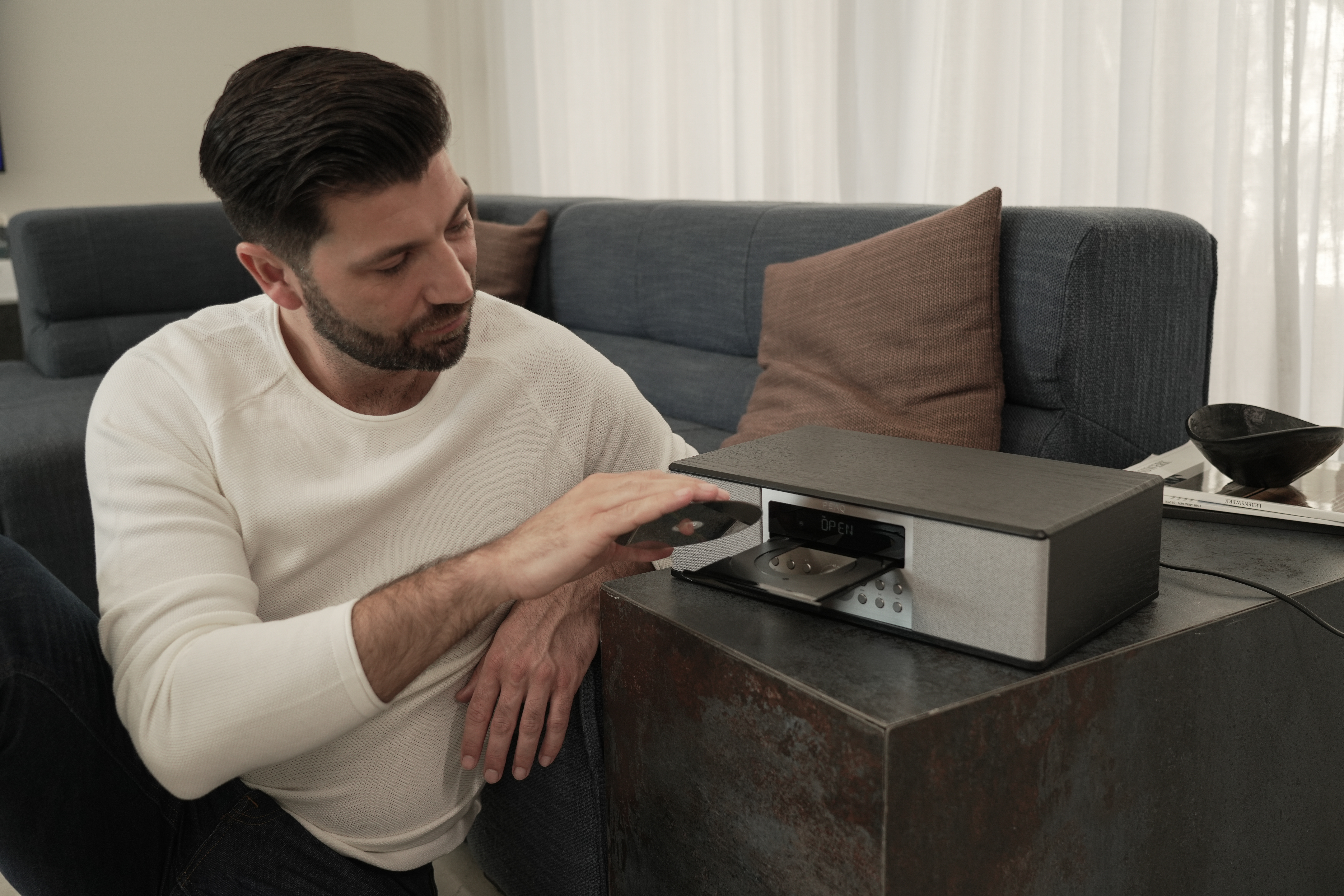 Mann, der eine CD in eine schwarz-silberne Stereoanlage einlegt, in der Nähe eines blauen Sofas und eines braunen Kissens.