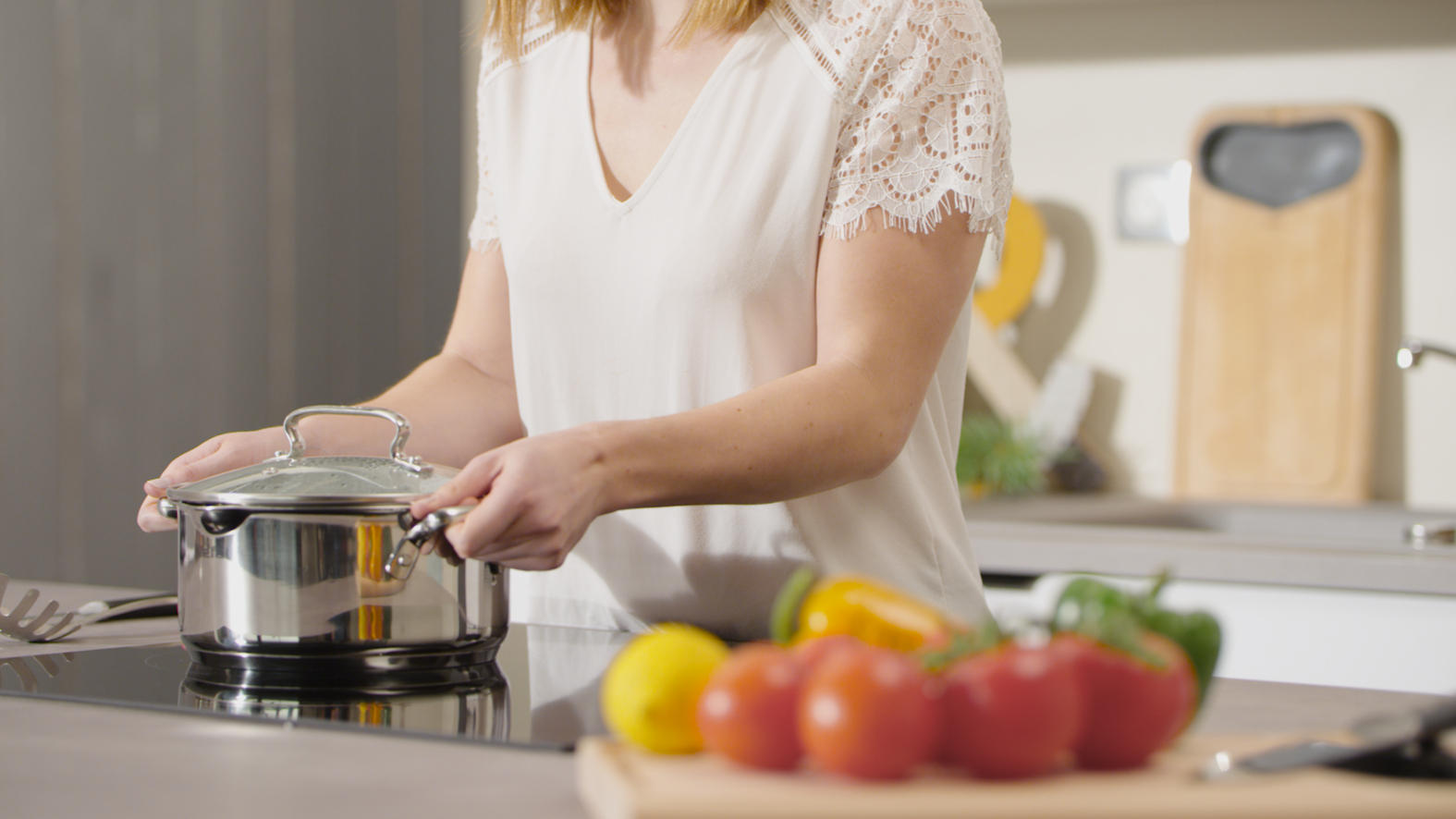 Kobieta w białej koszuli w kuchni, trzymająca garnek z pokrywką, w pobliżu warzyw.