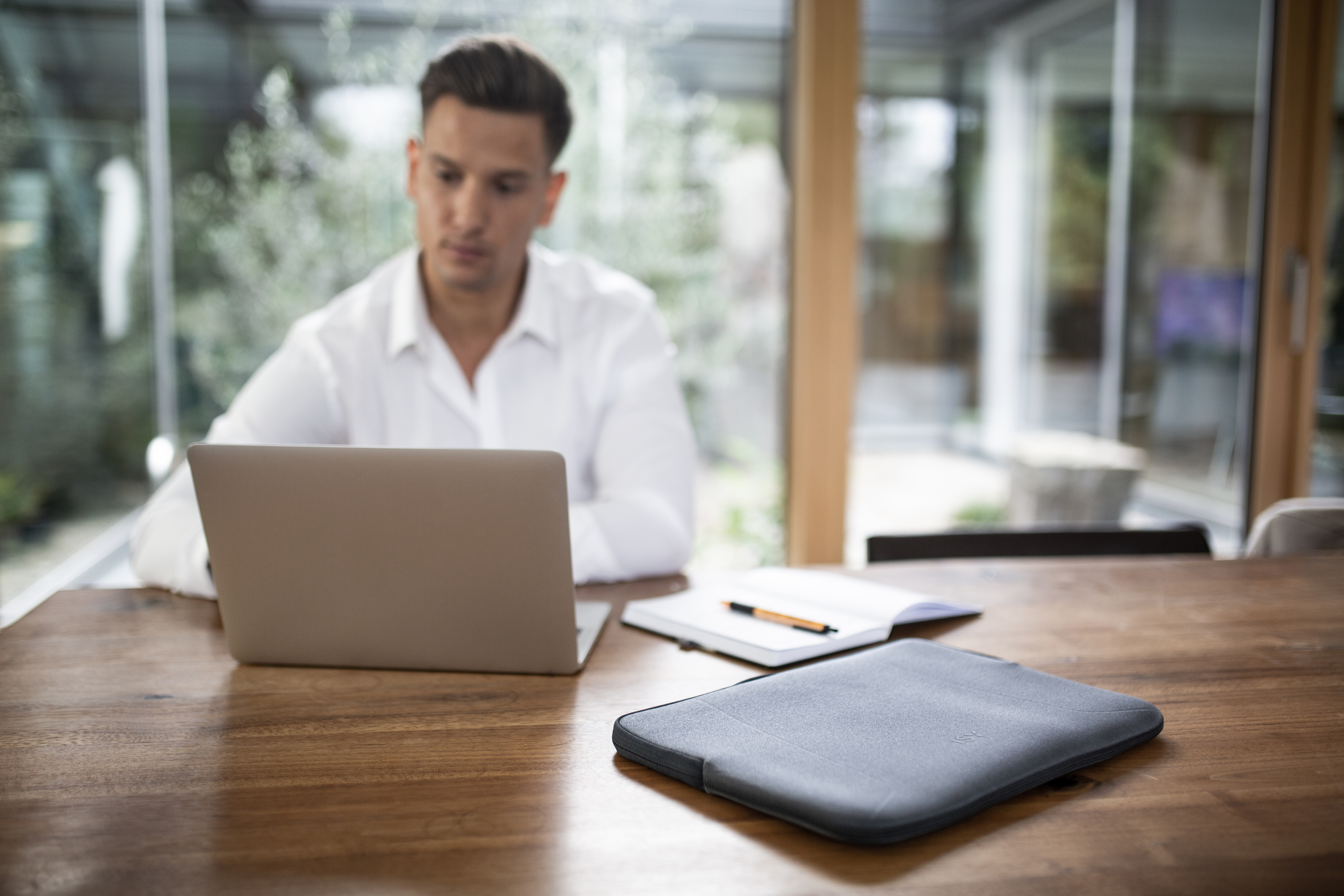 Mężczyzna siedzi przy drewnianym stole z laptopem, notatnikiem i etui. Tło rozmyte.