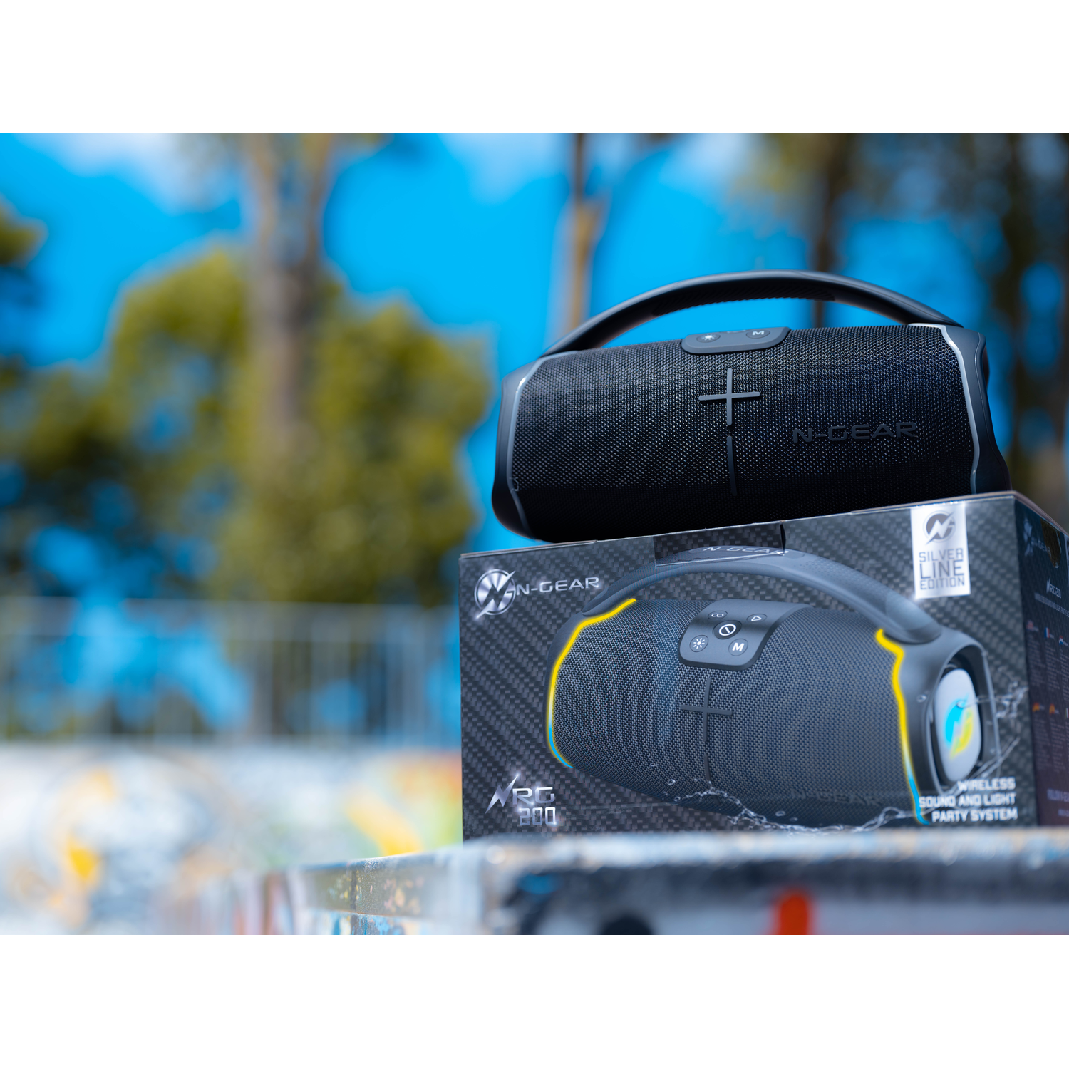 Black speaker with handle on top, sitting on a box with the same design, blue sky.