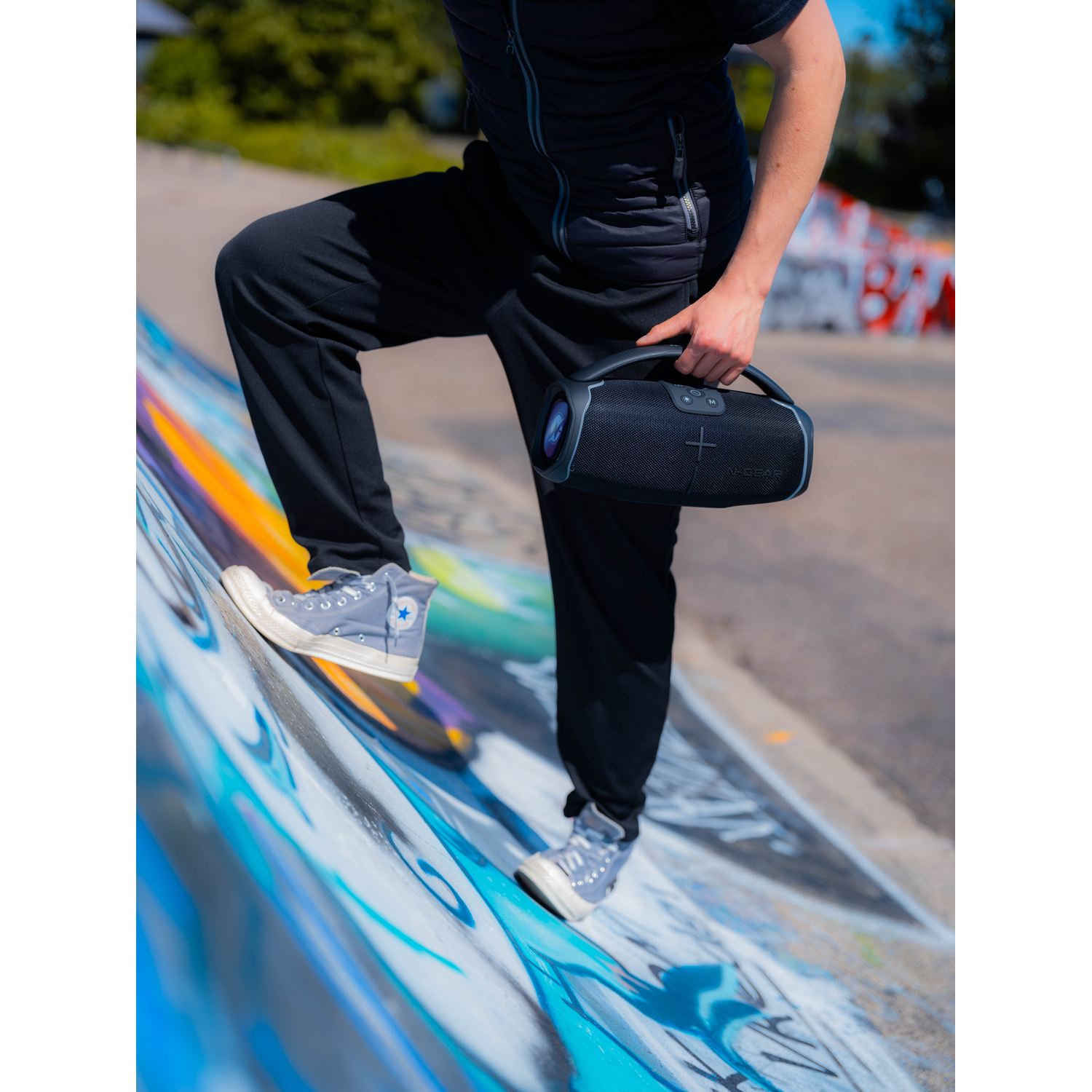 A person holding a black N-GEAR speaker, walking on a graffiti wall.