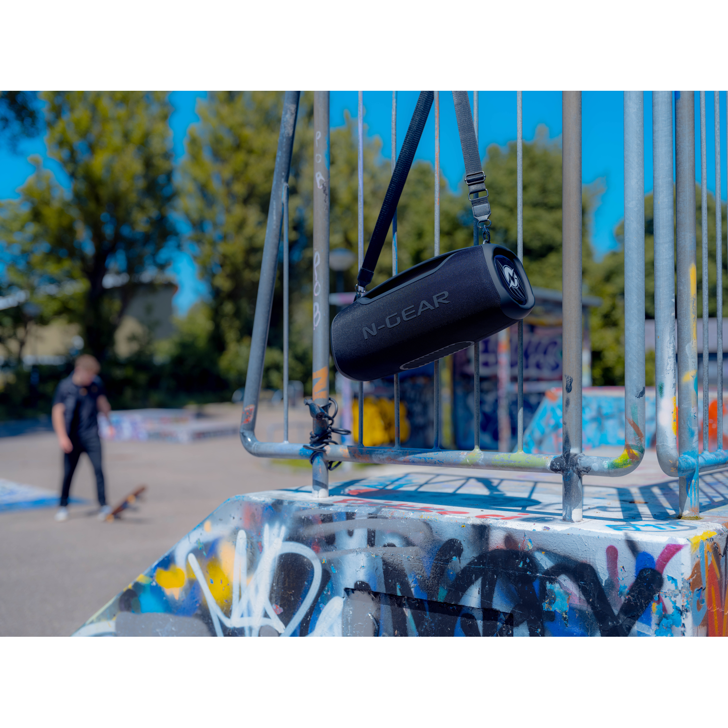 Black N-Gear speaker hanging on a rail, in a skate park. A person is skateboarding in background.