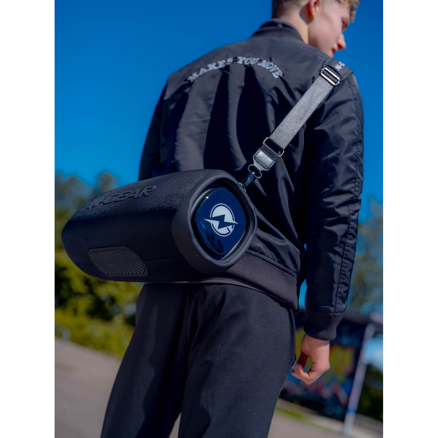 A man wearing a black jacket with a speaker bag slung over his shoulder against a blue sky.