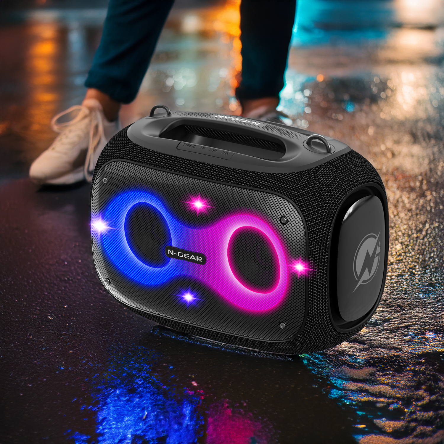Black speaker with blue and pink lights sits on a wet surface. A person's legs and shoes are in the background.