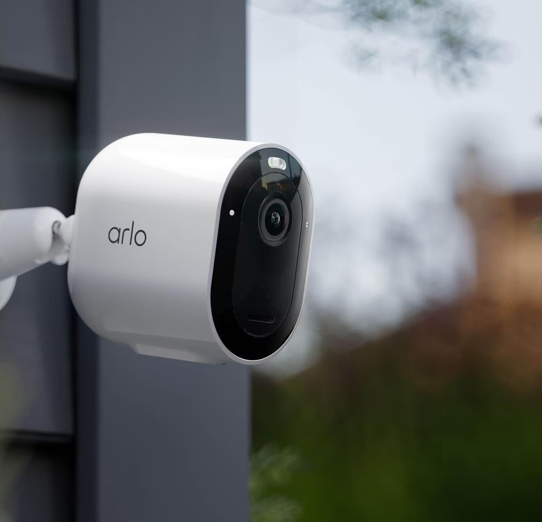 Une caméra de sécurité Arlo blanche montée sur une surface grise, avec un arrière-plan vert flou.
