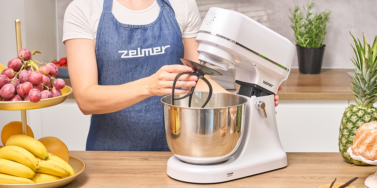 Kobieta używająca białego miksera stojącego, owoce i rośliny na blacie w tle.