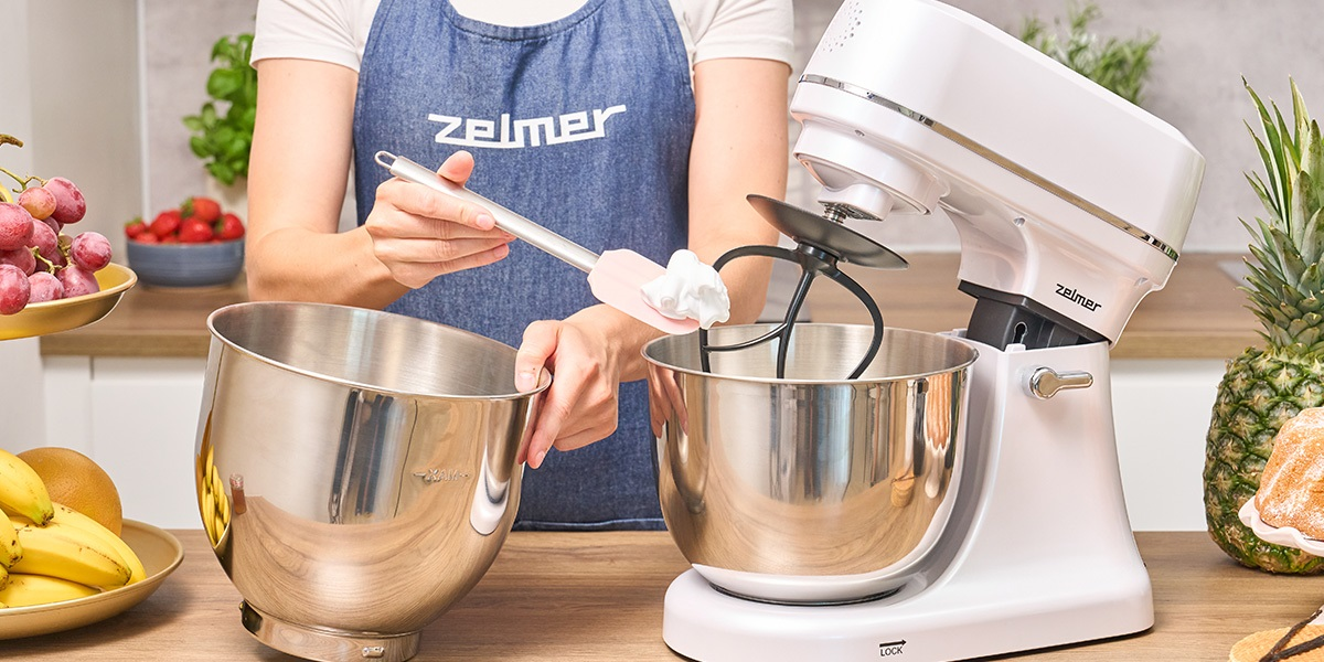 Kobieta używa szpatułki i miksera stojącego. Owoce, rośliny na blacie w tle.