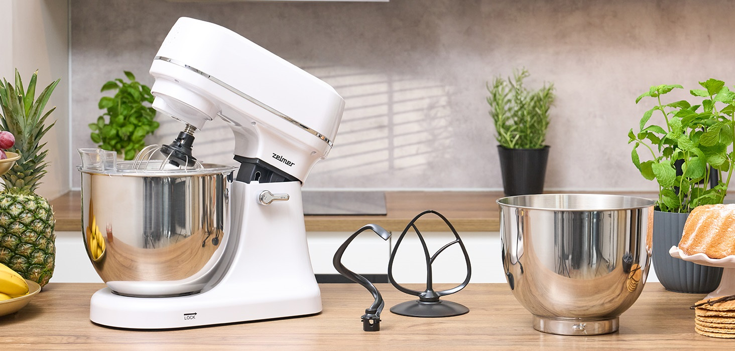 Biały mikser stojący z przystawkami. Rośliny, owoce i inne przedmioty na blacie.
