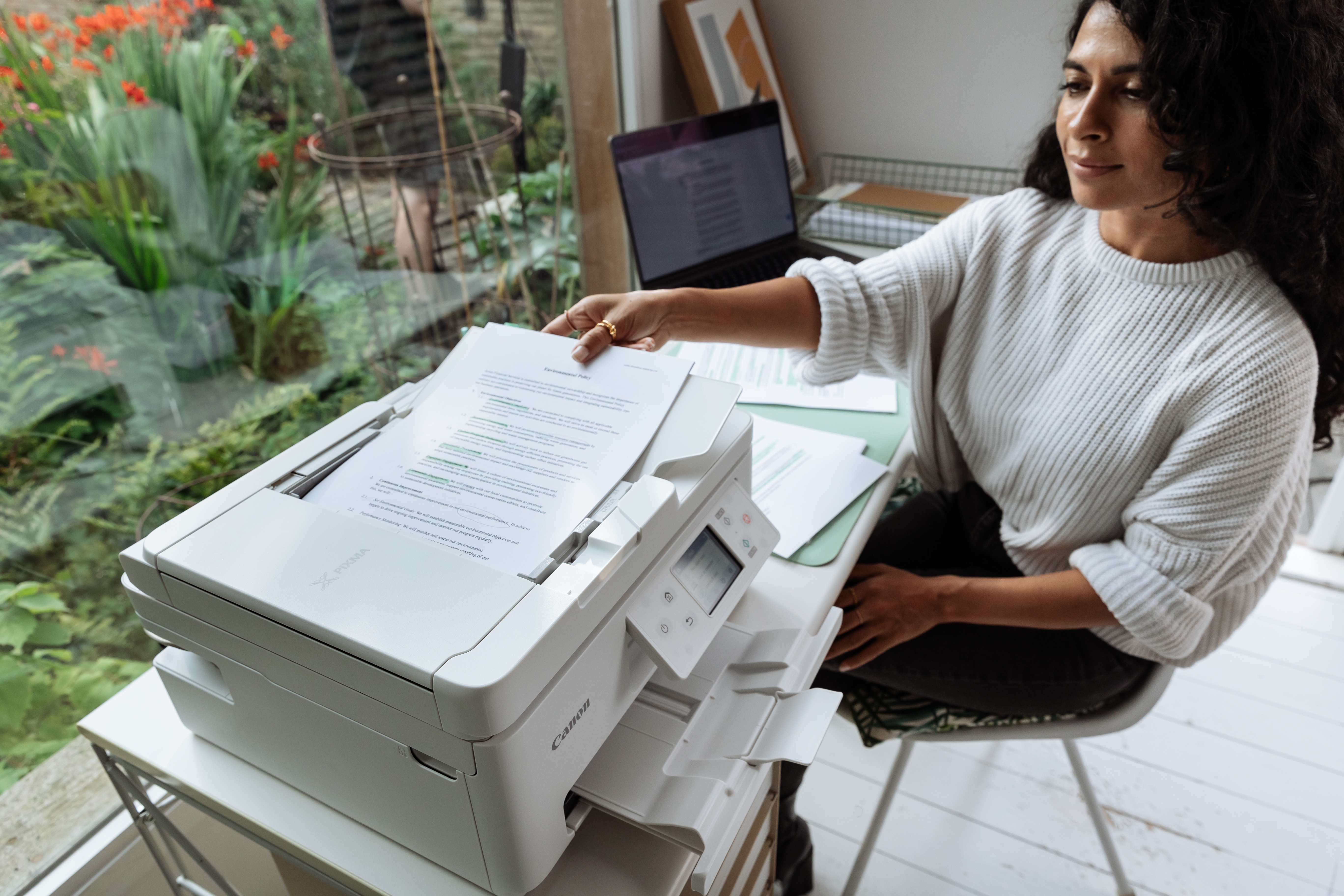 Eine Frau druckt an einem Schreibtisch Papiere mit einem weißen Drucker. Ein Laptop und Pflanzen sind in der Nähe.