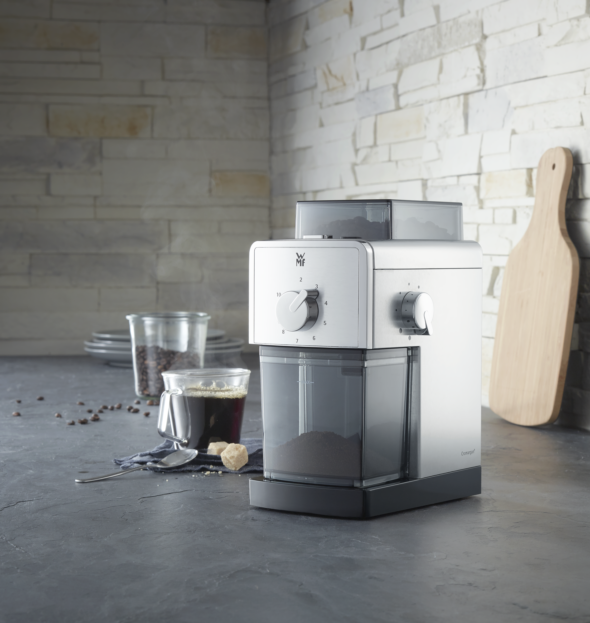 Silberne Kaffeemühle mit Kaffeepulver, Glas und Holzbrett auf einer Arbeitsplatte.