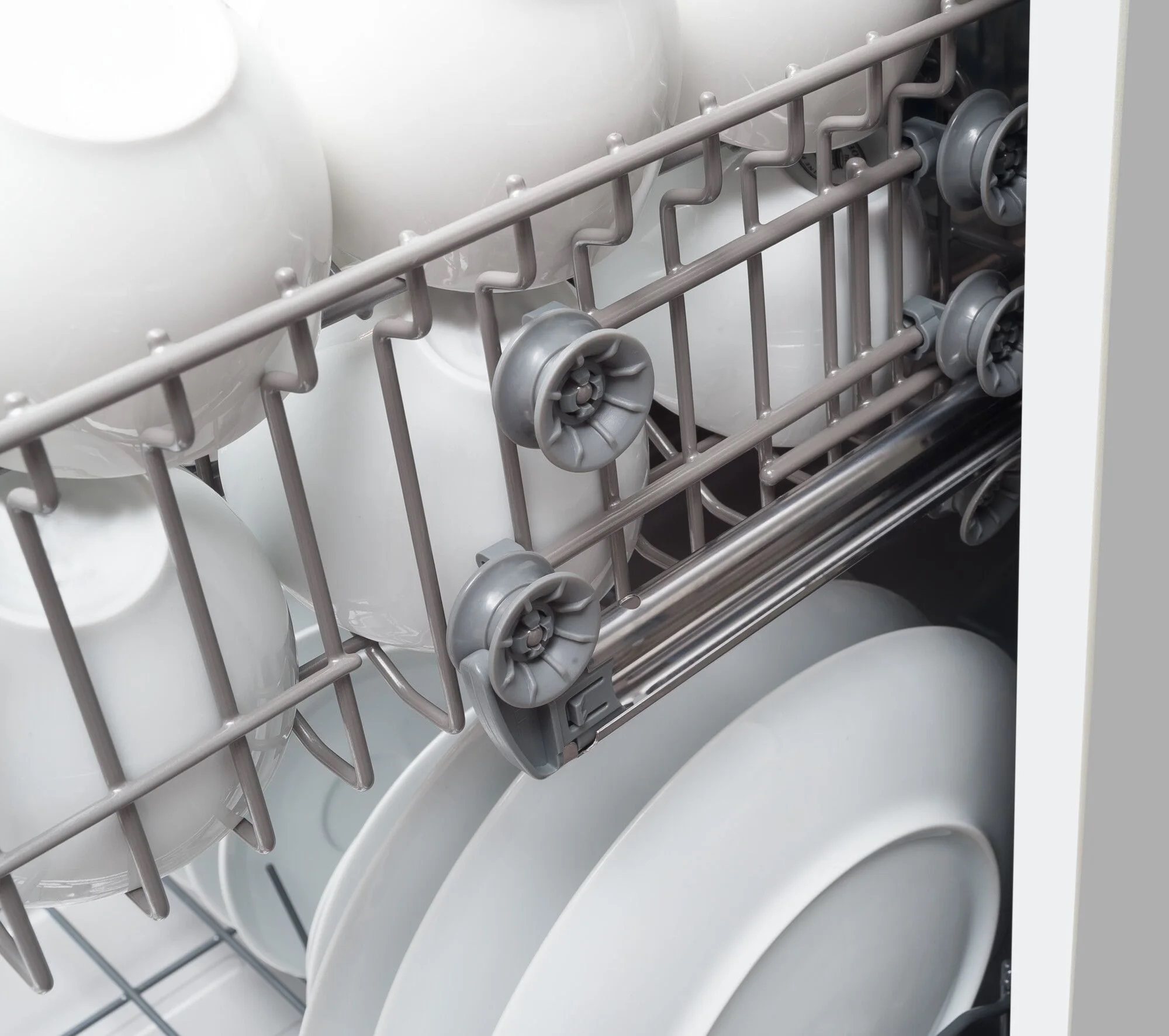 Górny kosz zmywarki z białymi naczyniami i małymi szarymi plastikowymi rolkami.