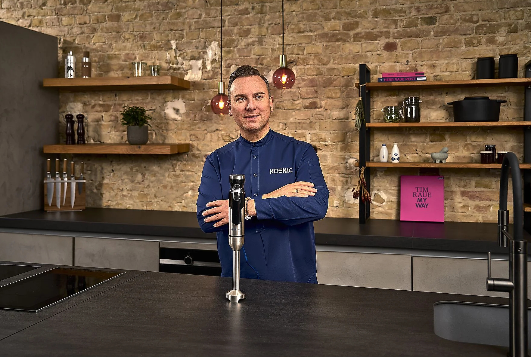 Mężczyzna w niebieskiej koszuli trzymający srebrny blender ręczny w nowoczesnej kuchni.