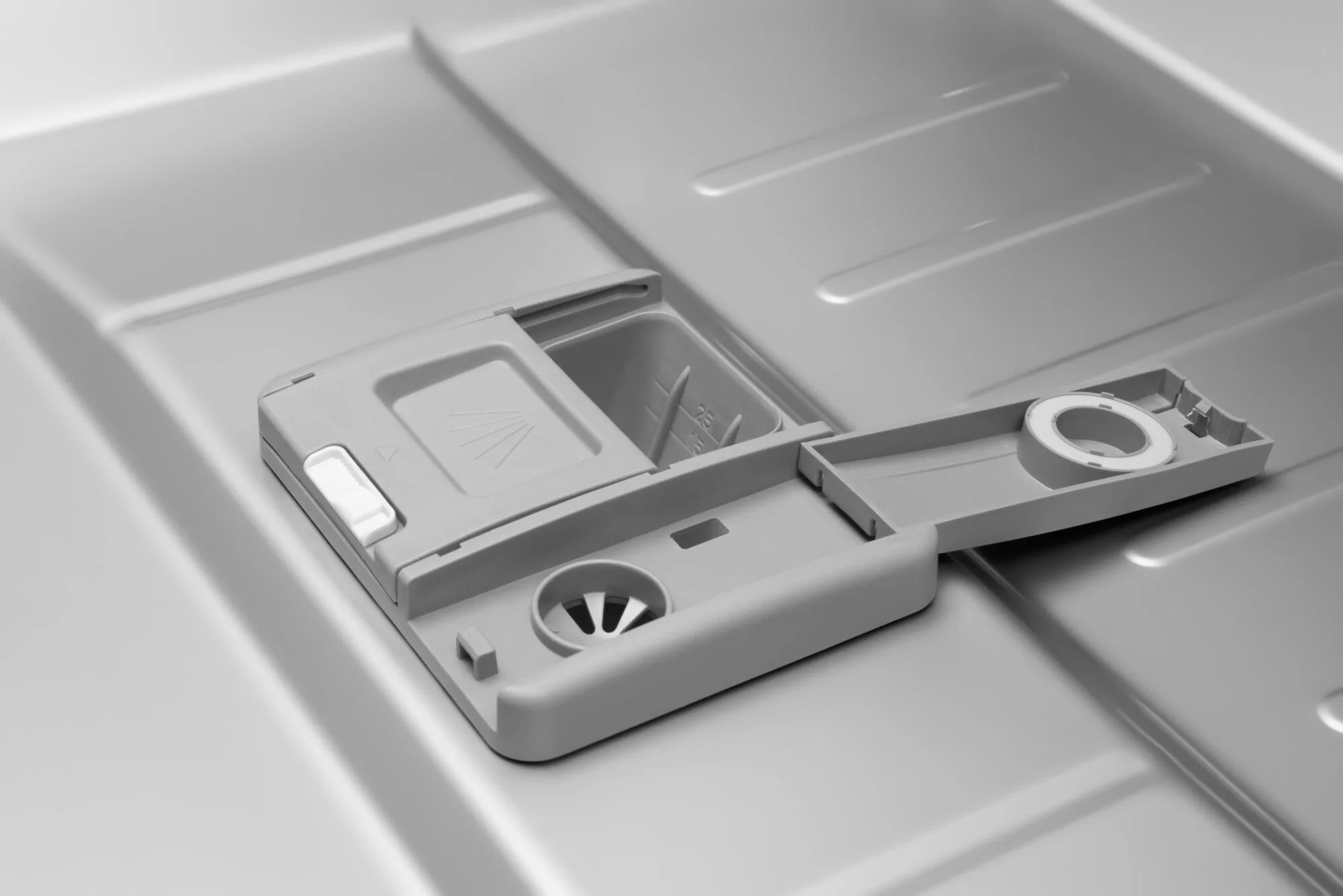 Szary dozownik detergentu do zmywarki, otwarty. Pokazuje przegródki i wzór natrysku.