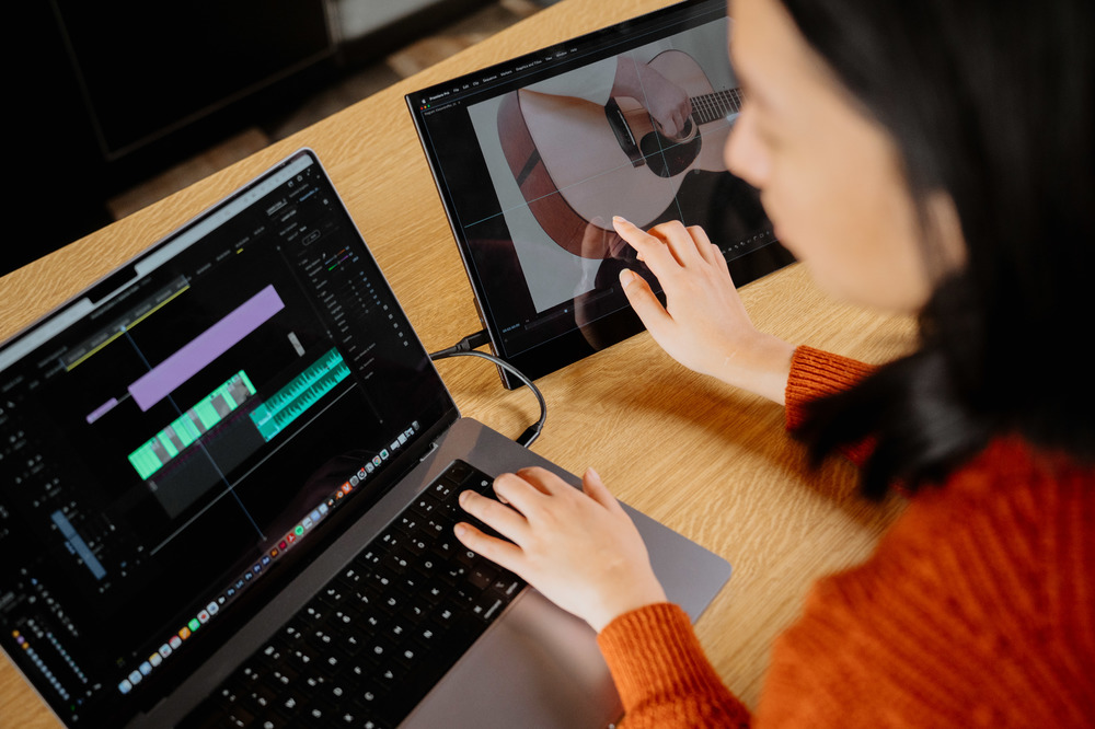 Donna che monta video su laptop e tablet. Mostra l'immagine di una chitarra sul tablet. Scrivania in legno, maglione arancione, capelli neri.