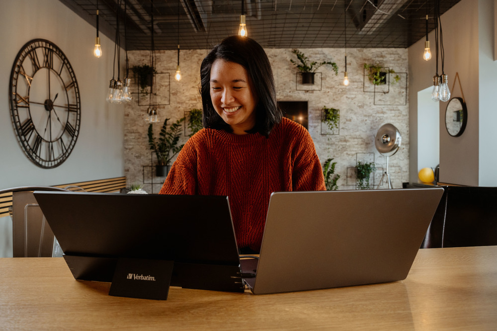 Una donna sorridente, che lavora su due laptop con una scheda Verbatim sul tavolo.