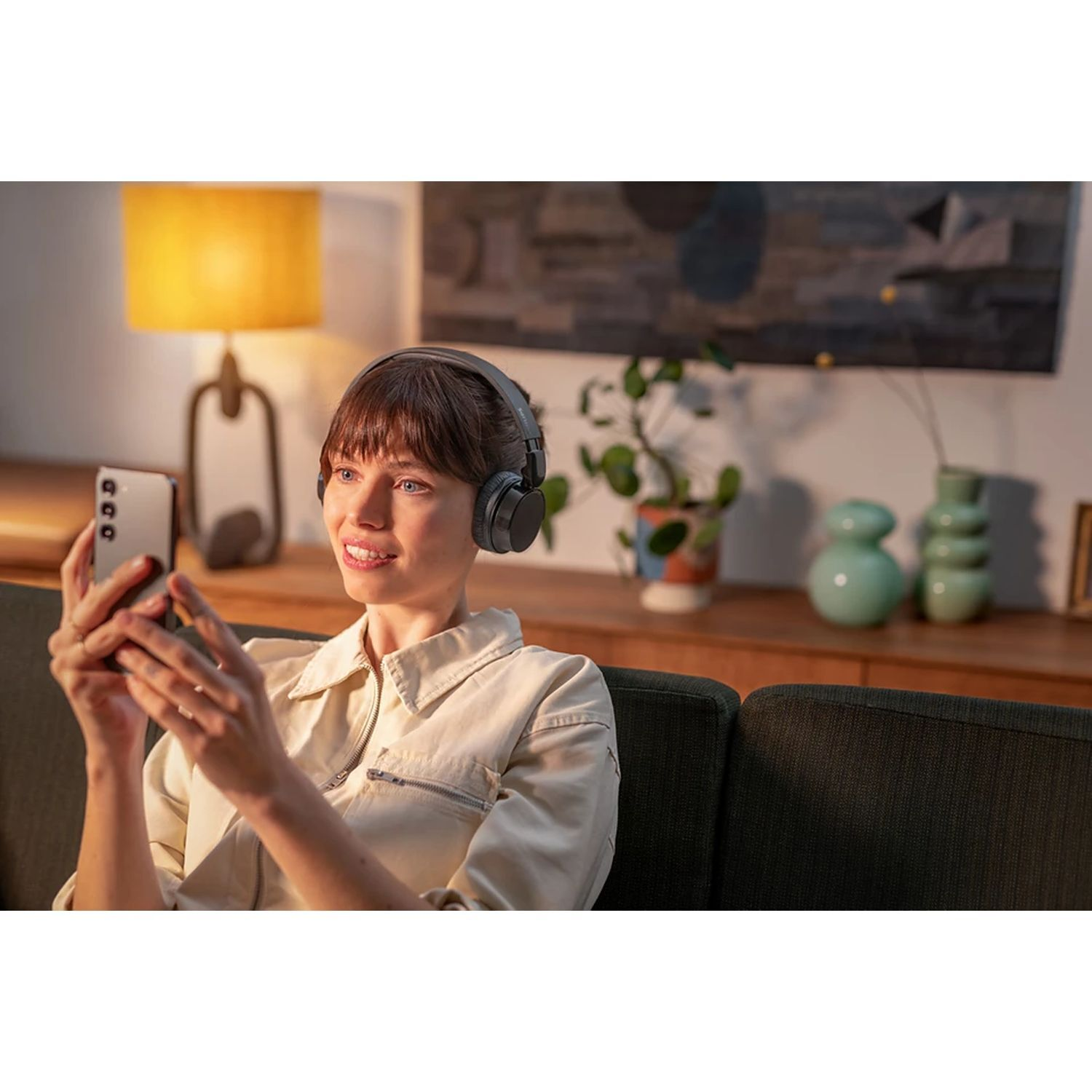 Femme avec casque, tenant un téléphone. Elle sourit en regardant le téléphone. Arrière-plan salon.