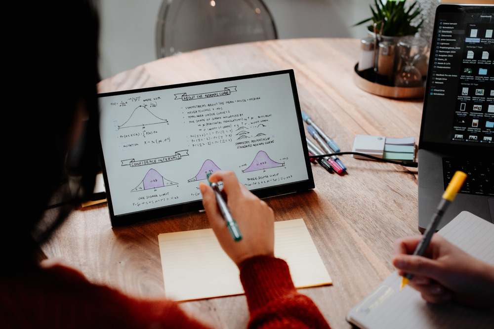 Persona che scrive su tablet con grafici matematici, altra persona che scrive su blocco note.