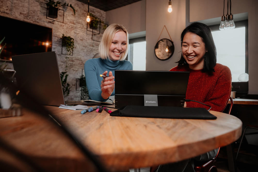 Due donne guardano un monitor, una indica. Tavolo con penne. Sfondo di un bar o ufficio.