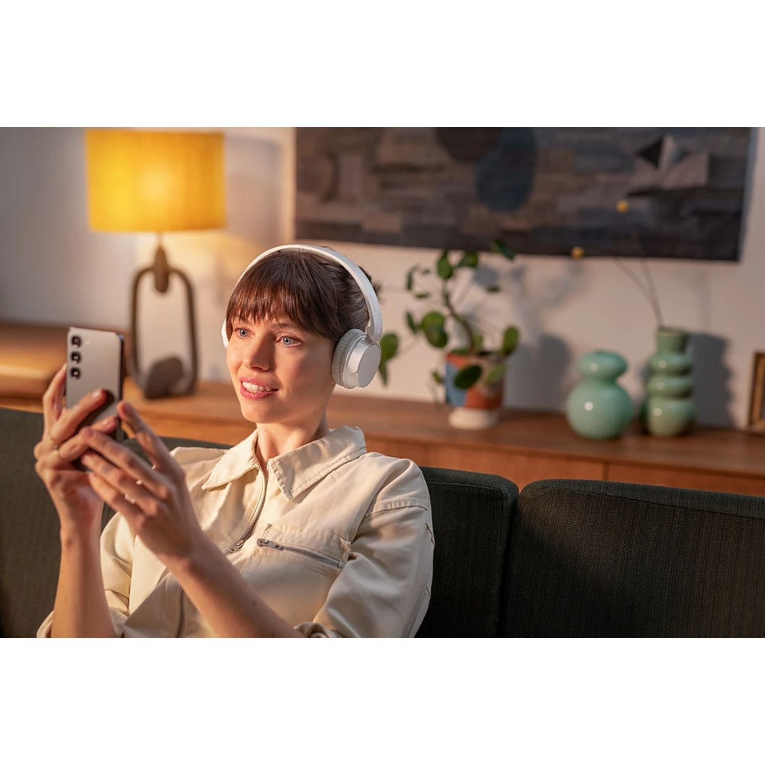 Woman with headphones and phone, smiling. Living room background, lamp, and furniture.